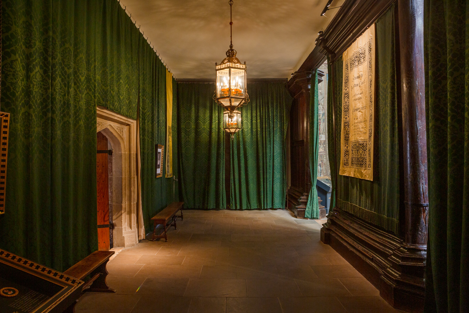 Hampton Court Palace. Photograph by Jayne Lloyd for Historic Royal Palaces. Photograph in Hampton Court Palace showing a dark room lit by two ornate brass and glass lanterns hanging from the ceiling in the middle, stone floor, the walls lined with patterned green curtains, a stone arched doorway to the left, wooden benches along the lefthand wall, a wooden wall to the right with a tapestry hanging in the centre and an opening at the far end into a daylit room.