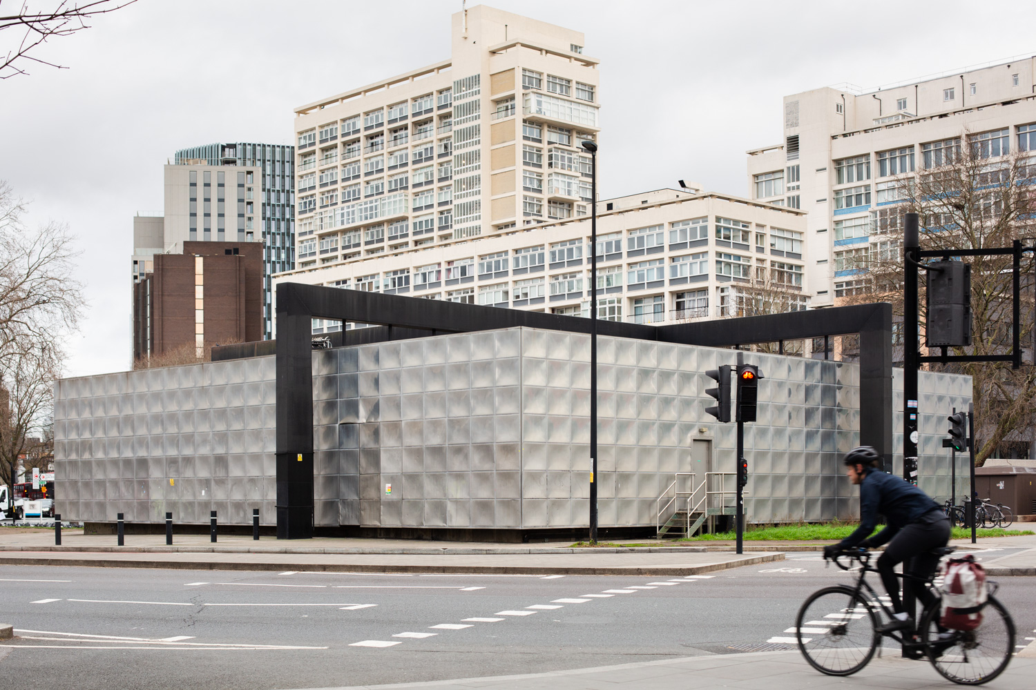 Photograph of the Michael Faraday Memorial, a large, squat metal building covered in indented squares of metal, with a black metal frame crossing over. In the distance tower blocks are visible and in the foreground is a road with a cyclist blurred as they pass.