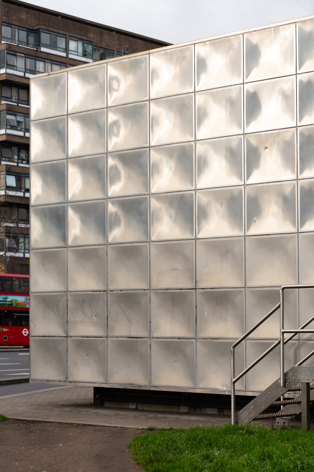 Photograph of a corner of the Michael Faraday Memorial, a large, squat metal building covered in indented squares of metal, catching the sunlight. There's a metal staircase to the right of the frame and a towerblock and red London bus can just be seen behind, to the left of the frame.