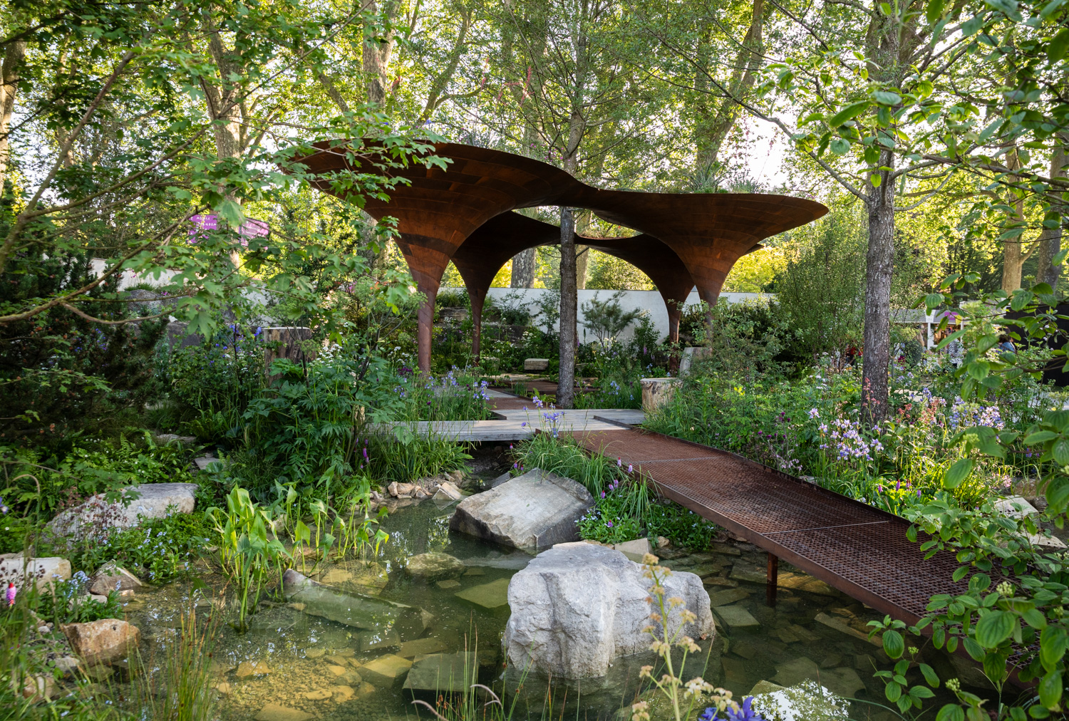 Photograph of a garden with large rusty-metal structures surrounding a tree towards the back centre of the frame, with a raised metal walkway leading towards it from the bottom right of frame. Lush green plants and flowers surround these, and in the foreground is a pool of water with stones and blocks of concrete in.