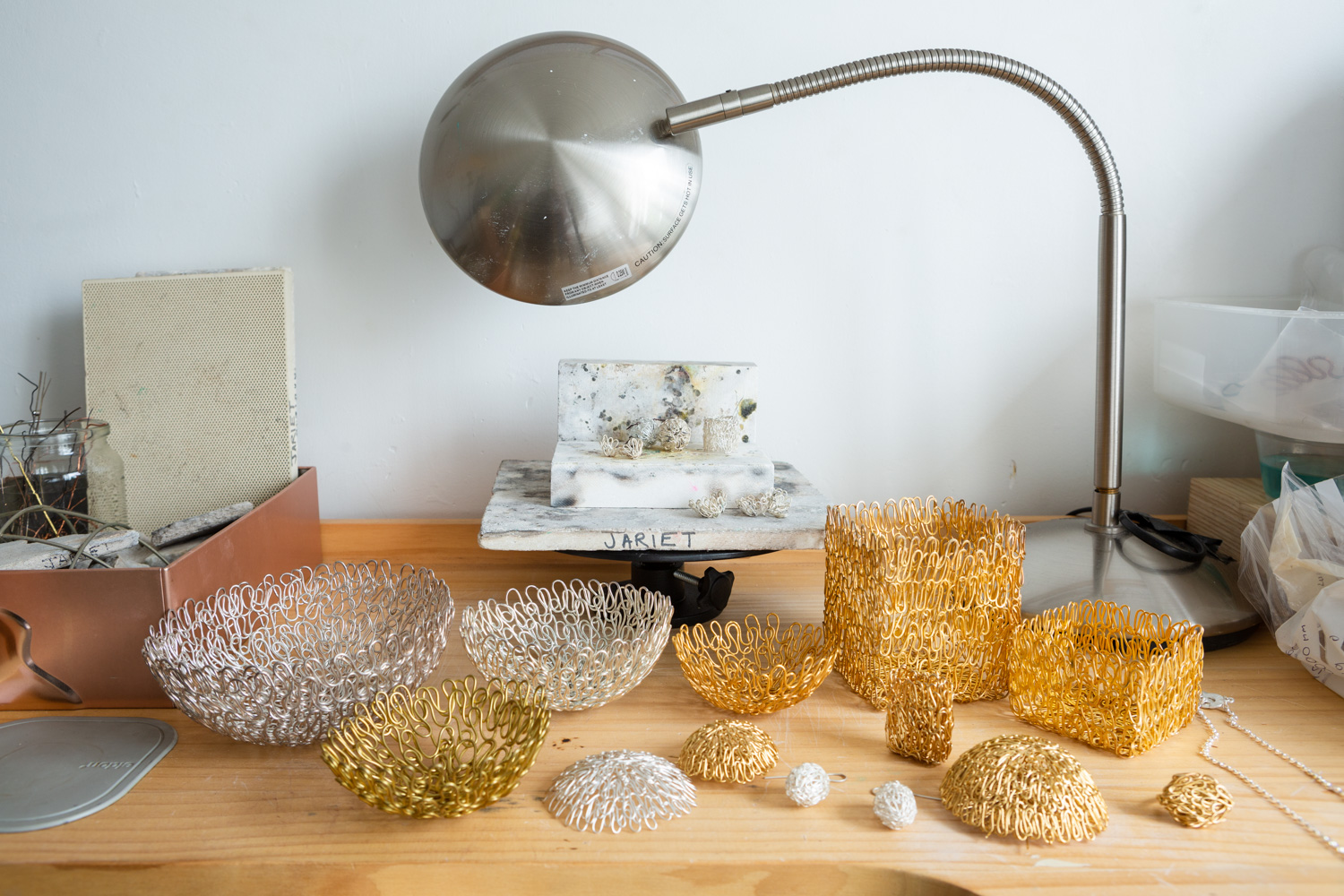 Artist Jariet Oloyé's studio, photographed by Jayne Lloyd for The Goldsmith's Company. Photograph of a wooden workbench with silver and gold pieces by Jariet Oloyé on display. Behind is a tile on a raised platform with 'JARIET' handwritten on it, there's a metal table lamp behind and other objects are visible just out of shot to the left and right.