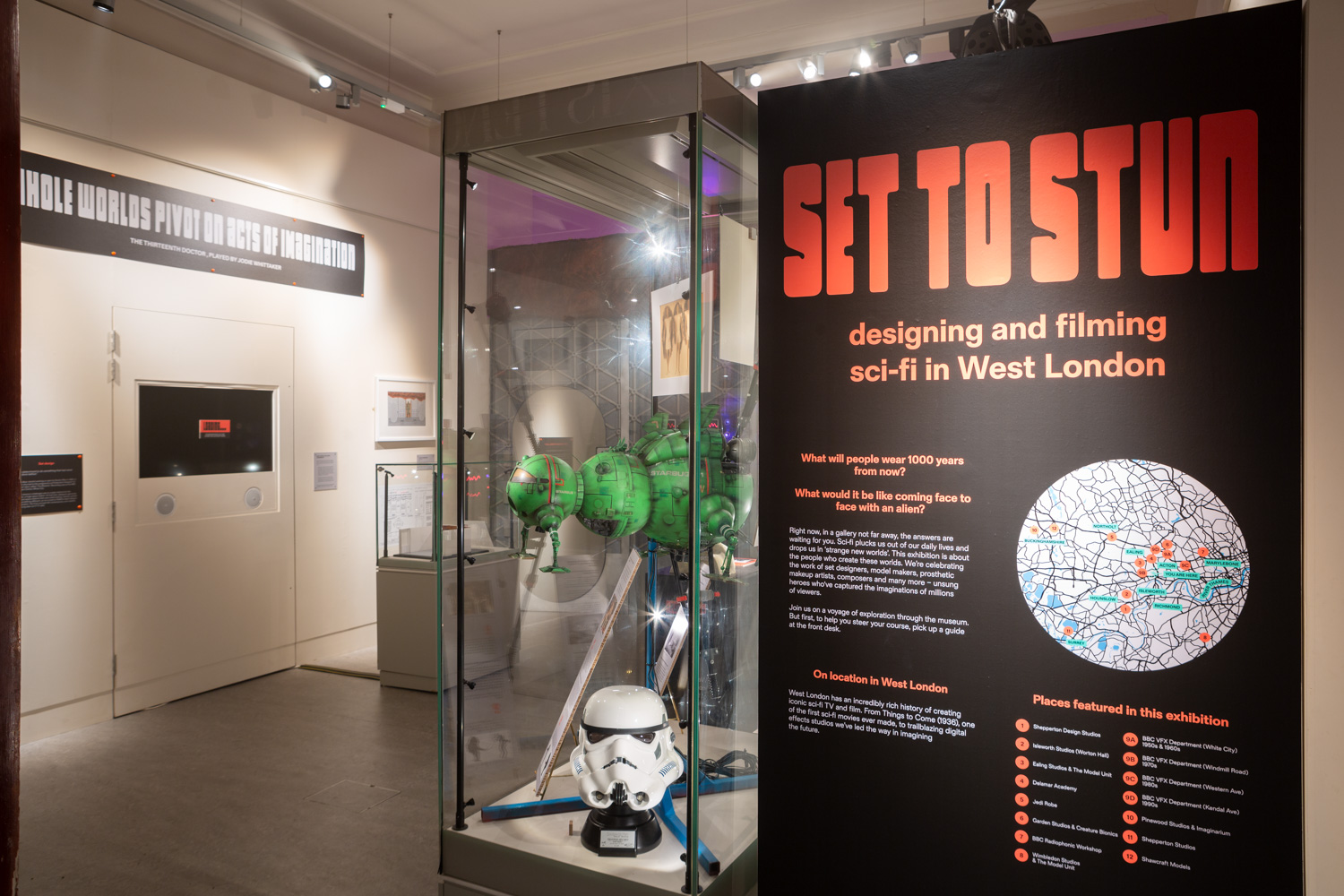 Set to Stun at Gunnersbury Park Museum. Photograph by Jayne Lloyd. Photograph of 'Set to Stun' at Gunnersbury Park Museum showing an information board to the right saying 'Set to Stun, designing and filming sci-fi in West London' with further text and a map in a circle below, a display case with items including a Stormtrooper helmet in the centre of the frame and further signs on the wall to the left including one saying 'Whole worlds pivot on acts of imagination'.