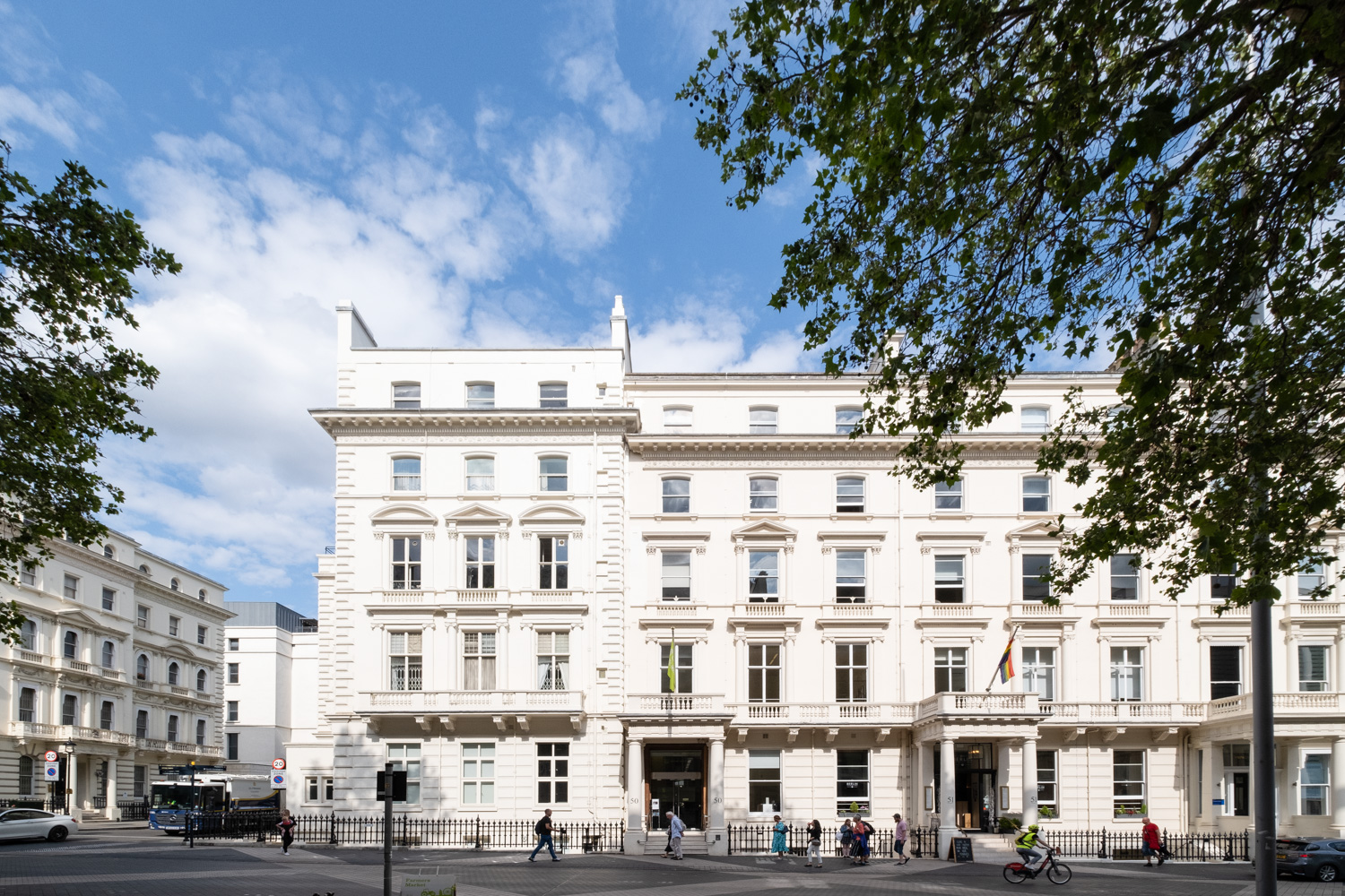 © Goethe-Institut London. Photo: Jayne Lloyd. Photograph of the Goethe-Institut London, a large white building with lots of windows and a columned entrance, blue sky with a few clouds above, people walking past on the pavement and leaves from trees in the foreground.