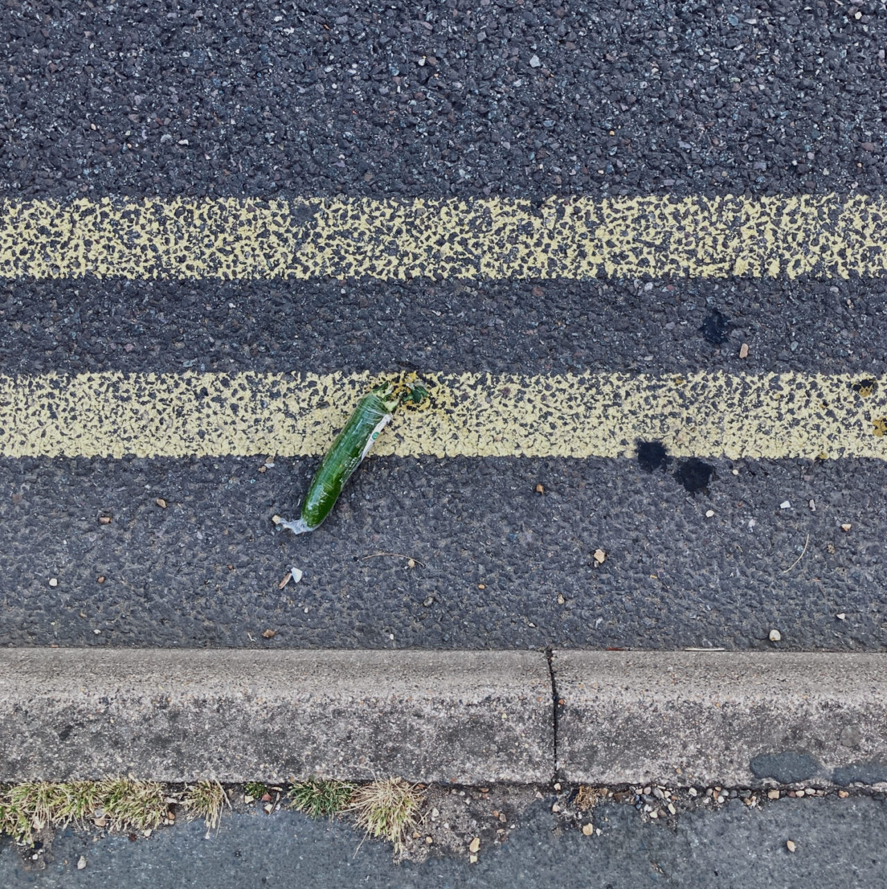 Photograph looking down at a cucumber in a gutter by the side of the road, with pavement at the bottom and double yellow lines above.