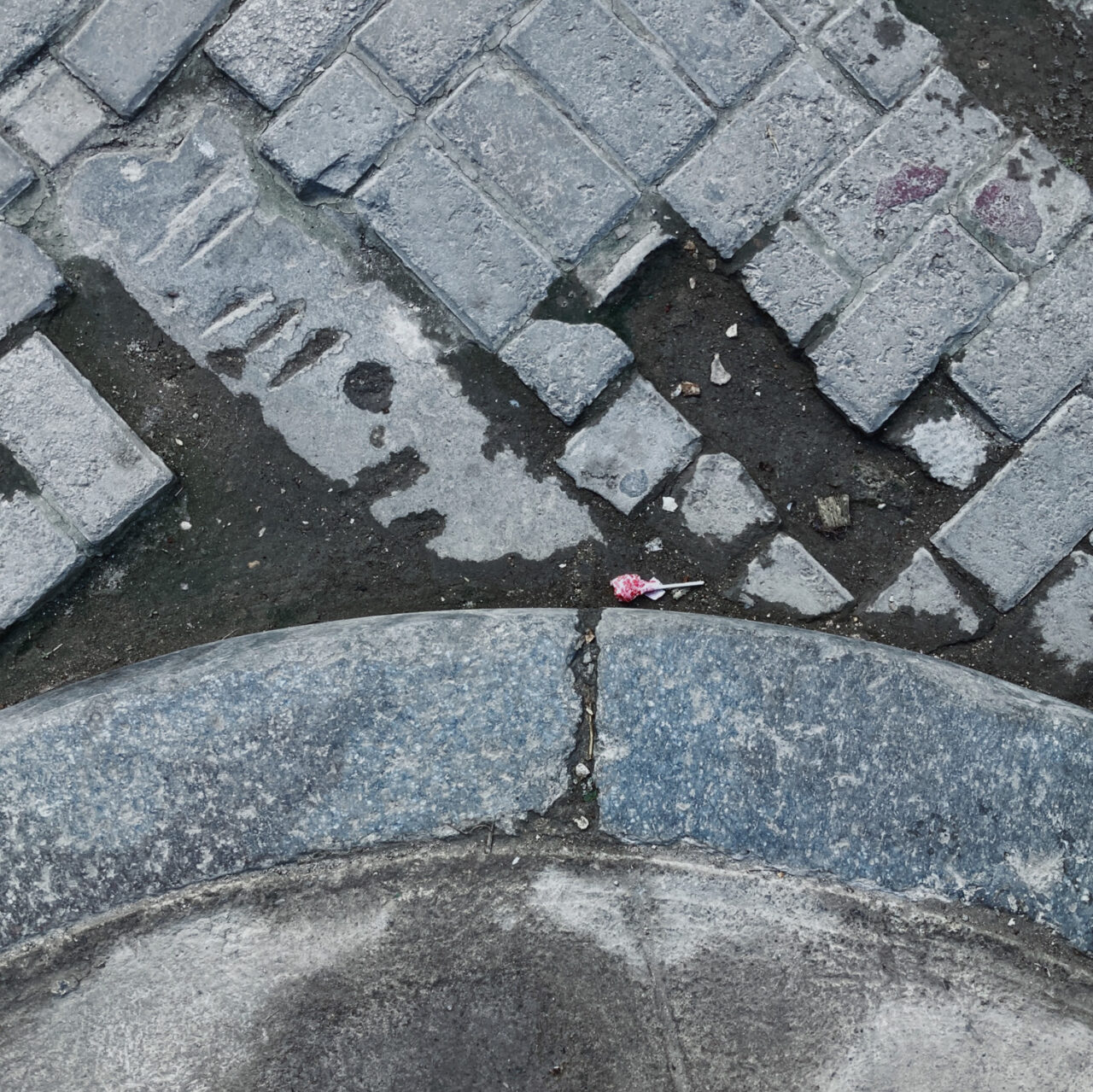 Photograph looking down at a lolly po in a gutter by the side of the road, with a corner of pavement at the bottom and tiles above.