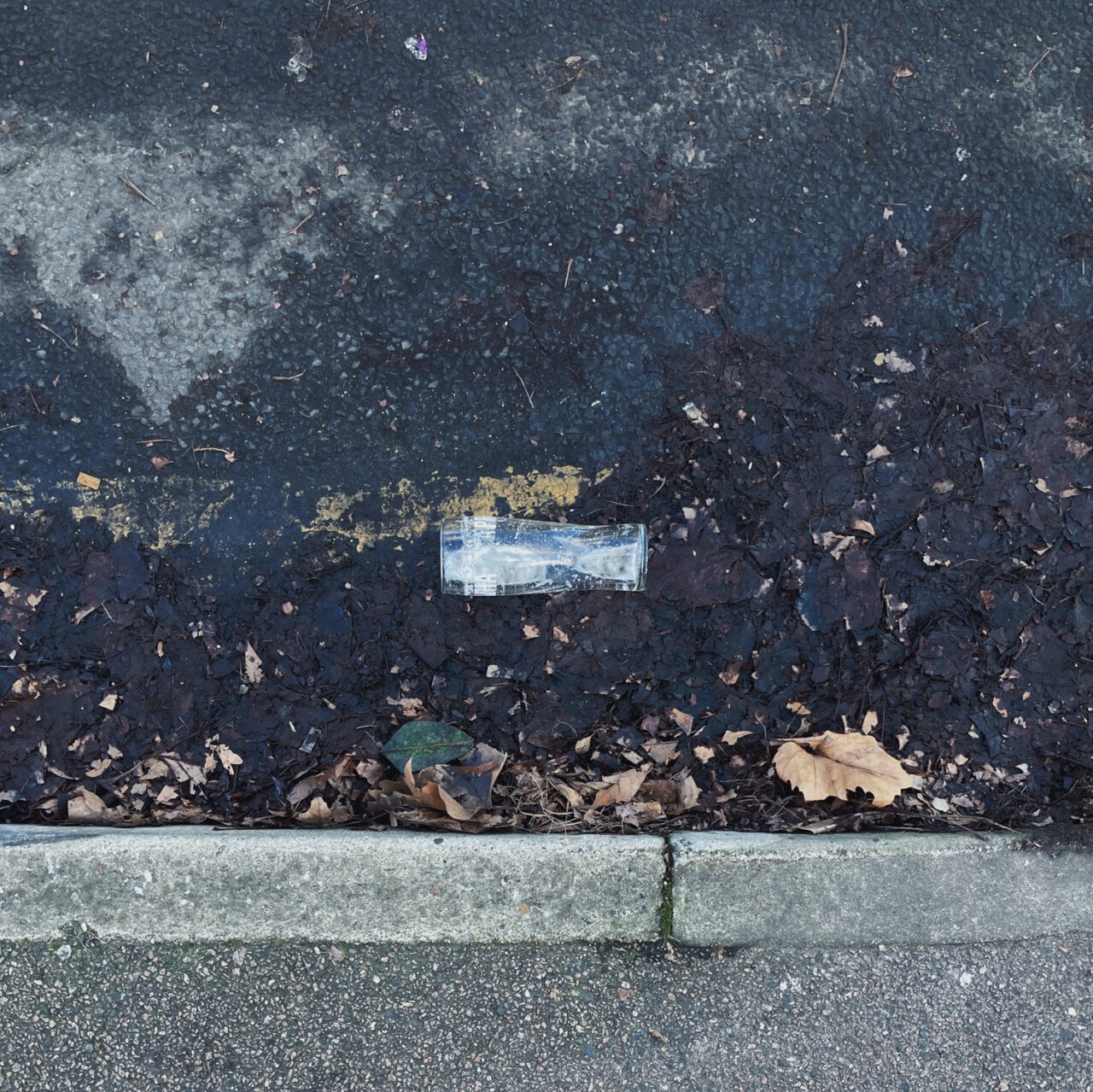 Photograph looking down at a pint glass on its side in a gutter by the side of the road, with pavement at the bottom and double yellow lines above.