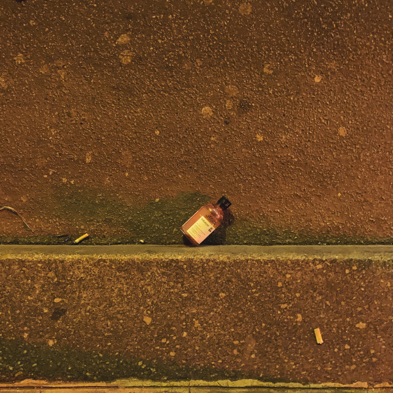 Photograph looking down at a bottle of liquid soap in a gutter by the side of the road, with pavement at the bottom and double yellow lines above.