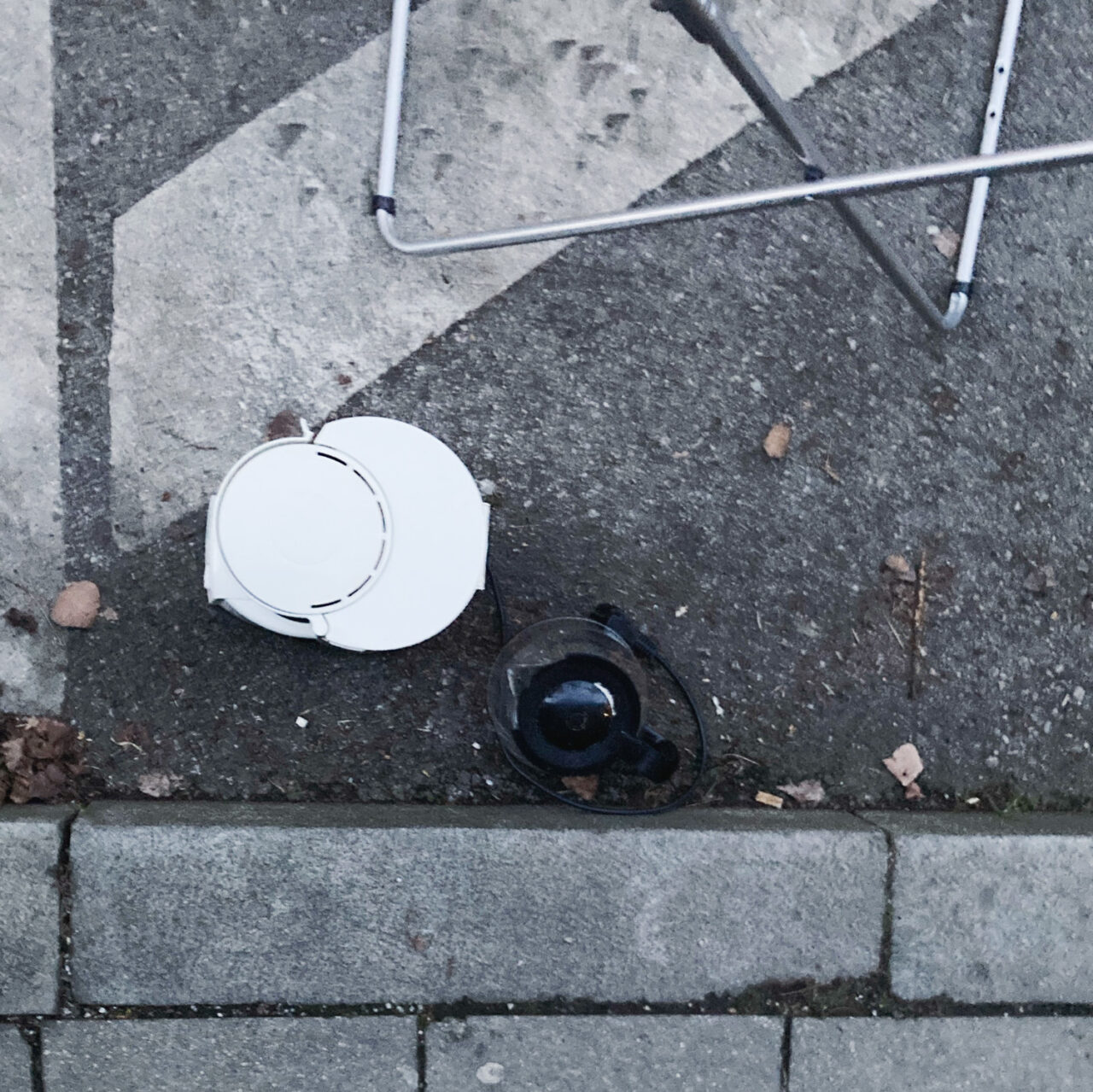 Photograph looking down at a filter coffee machine and jug in a gutter by the side of the road, with pavement at the bottom and double yellow lines above.