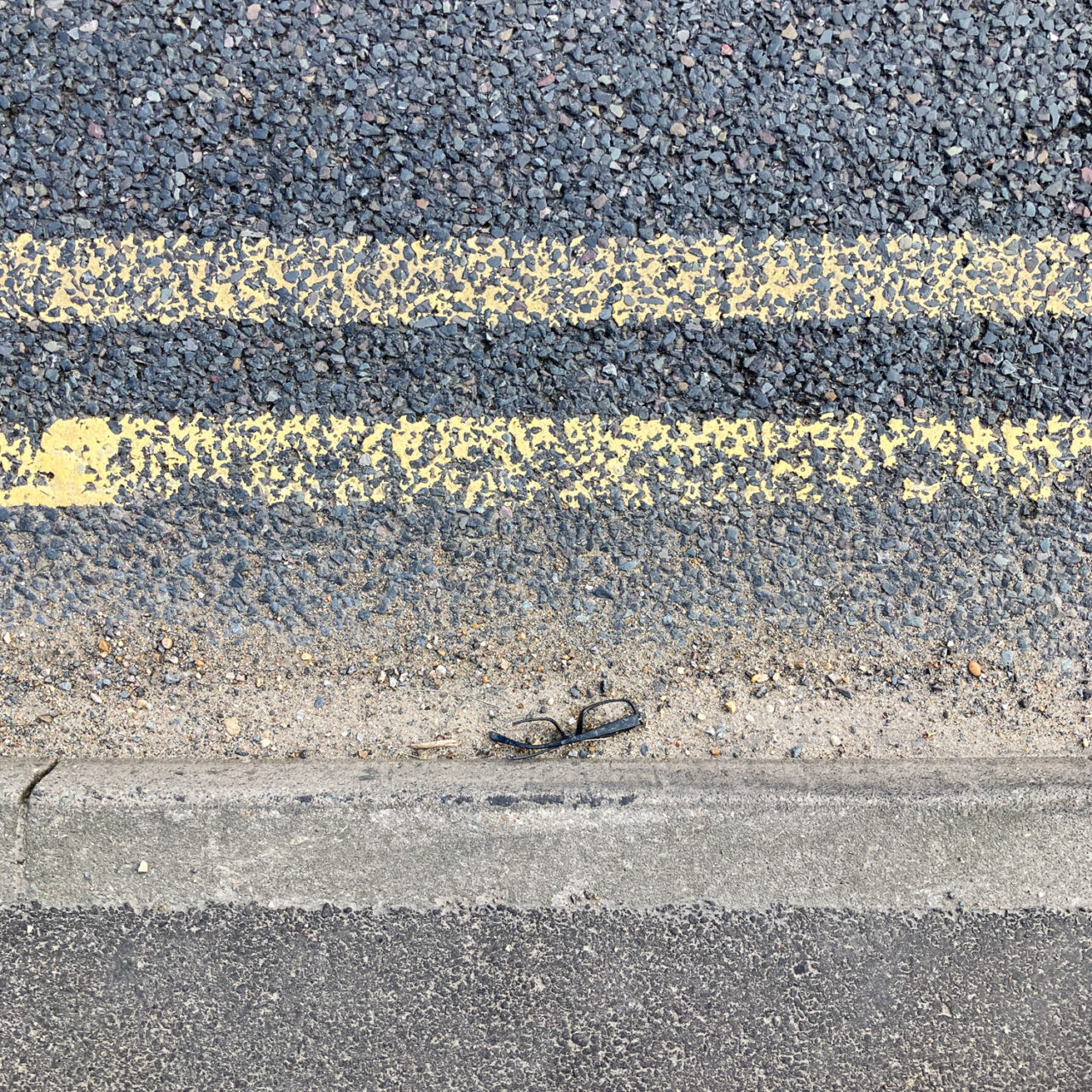 Photograph looking down at a pair of broken glasses in a gutter by the side of the road, with pavement at the bottom and double yellow lines above.