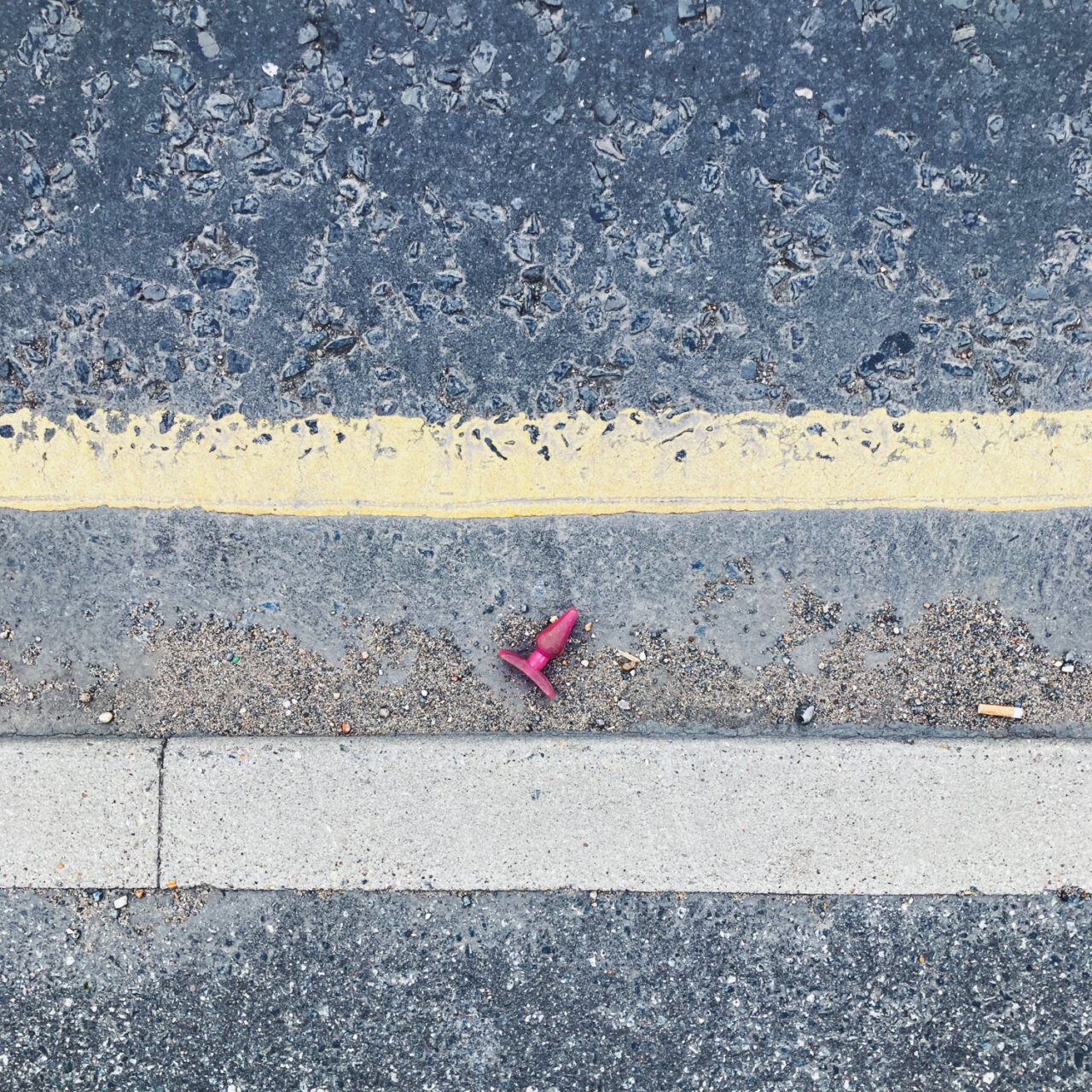 Photograph looking down at a pink adult toy in a gutter by the side of the road, with pavement at the bottom and single yellow line above.