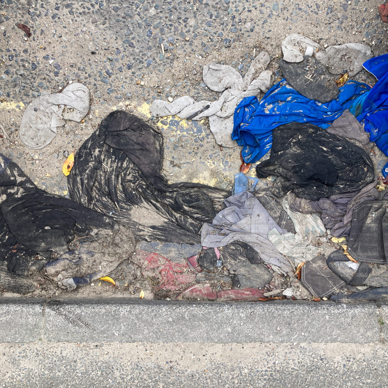 Photograph looking down at load of squashed clothes by the side of the road, with pavement at the bottom and double yellow lines barely visible beneath, all covered in dirt.