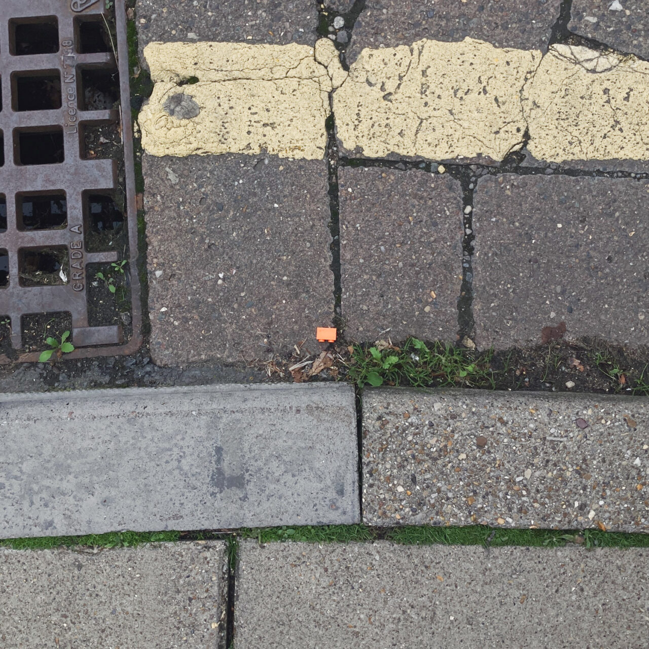 Photograph looking down at the gutter at the side of the road with the curb at the bottom of the frame, a painted yellow line at the top of the frame and a two-dot orange LEGO brick in the centre of the frame.