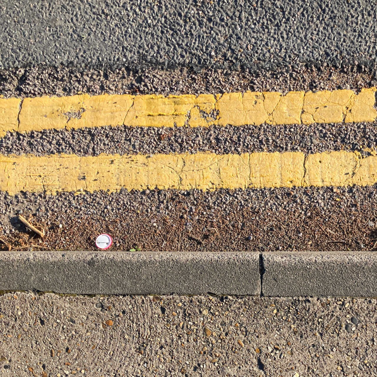 Photograph looking down at the gutter at the side of the road with the curb at the bottom of the frame, painted double yellow lines at the top of the frame and an upside down badge in the centre left of the frame.