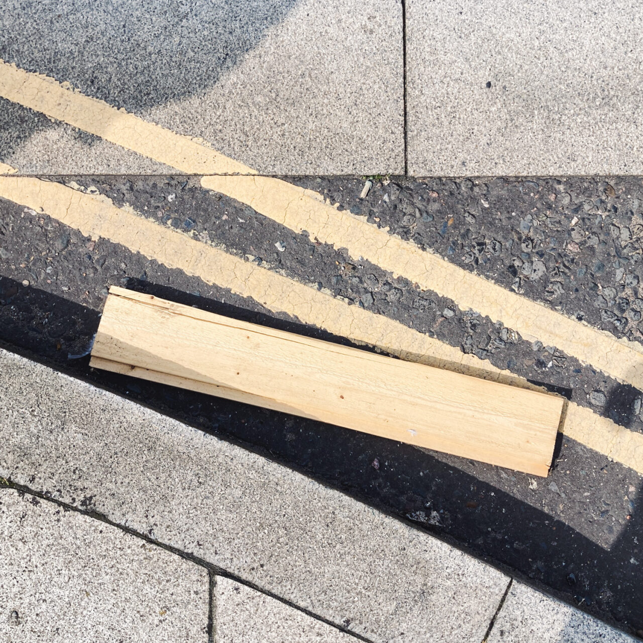 Photograph looking down at the gutter at the side of the road with the curb at the bottom left of the frame, a painted double yellow line in the centre of the frame with a plank of wood on top.