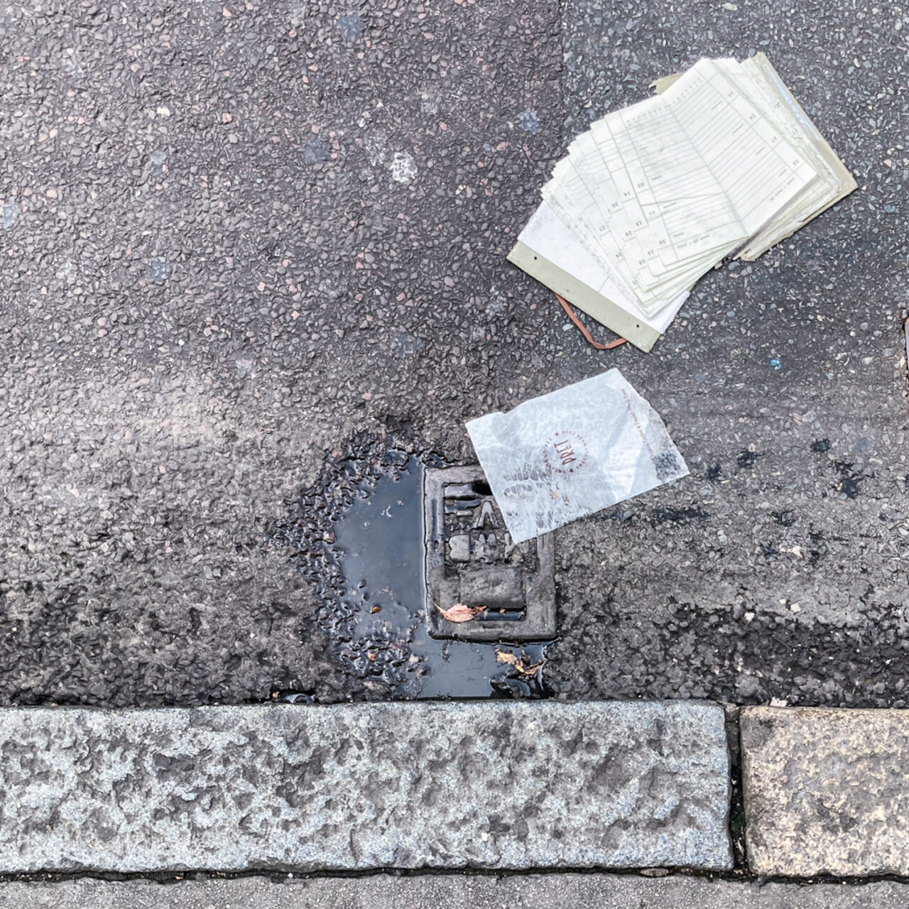 Photograph looking down at the gutter at the side of the road with the curb at the bottom of the frame and a flatterned diary at the top right of the frame, a puddle, utility cover and 'Pret' napkin towards the centre of the frame.