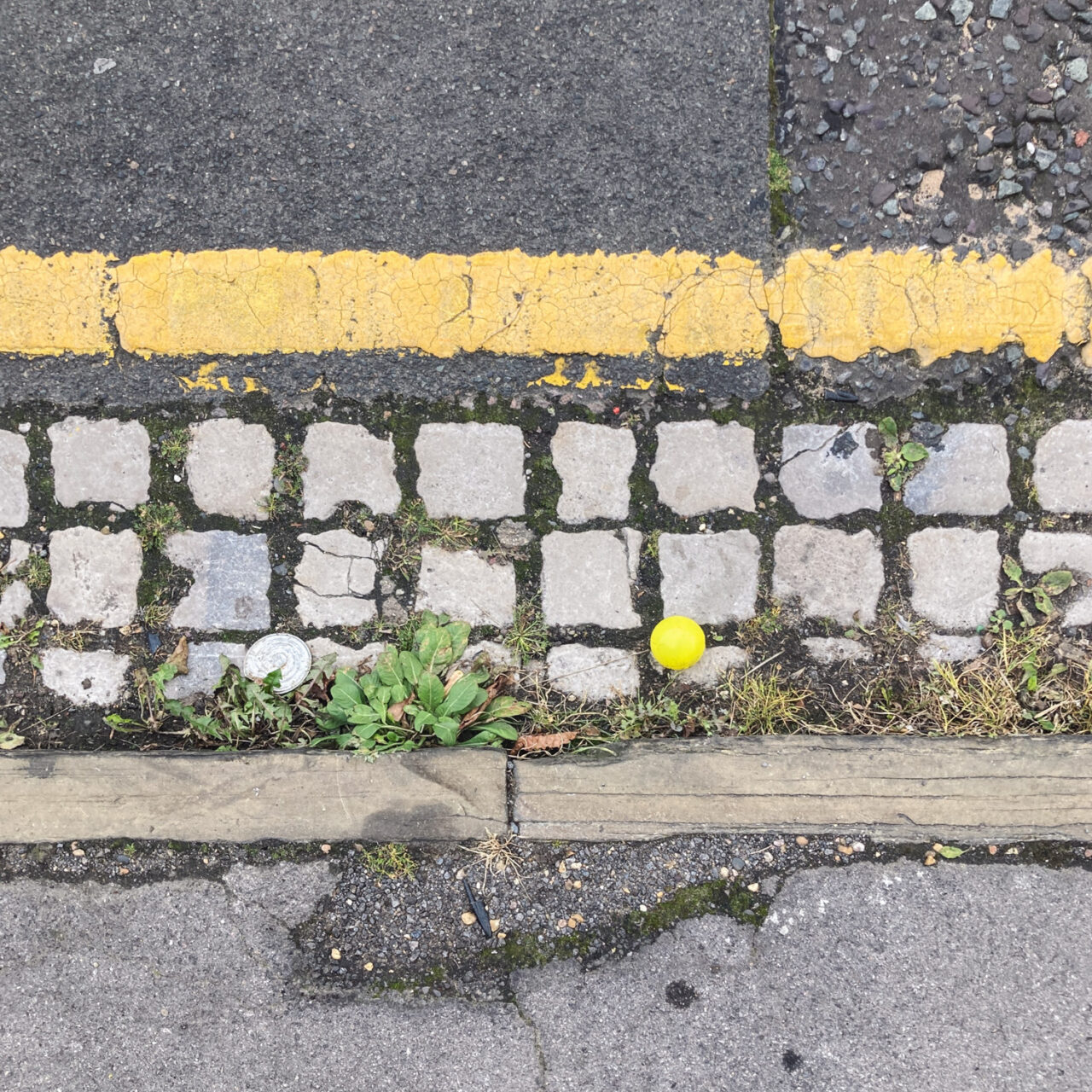 Photograph looking down at the gutter at the side of the road with the curb at the bottom of the frame, a painted yellow line at the top of the frame, weeds, cobble stones and a bright yellow plastic ball in the centre of the frame.