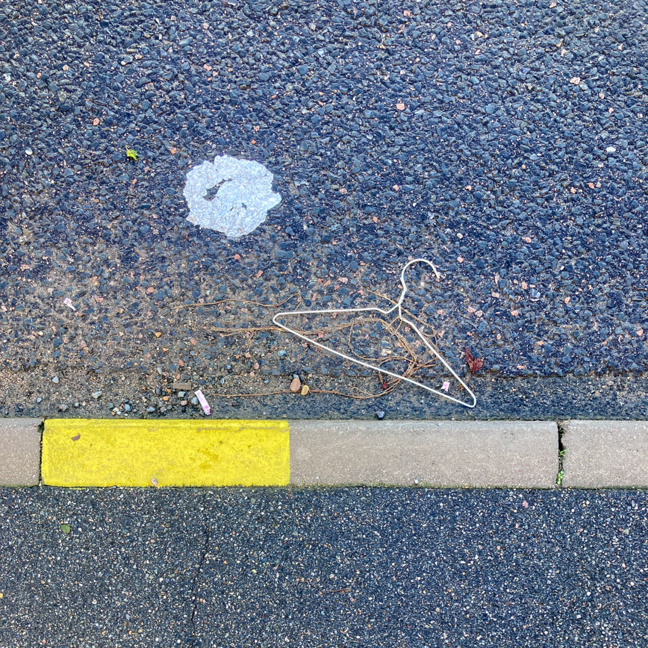 Photograph looking down a gutter at the side of a road, with a wire coat hanger in the centre of the frame and the curb below, with a rectangle to the left painted yellow. There is dirt and a big blog of something off-white in the road.