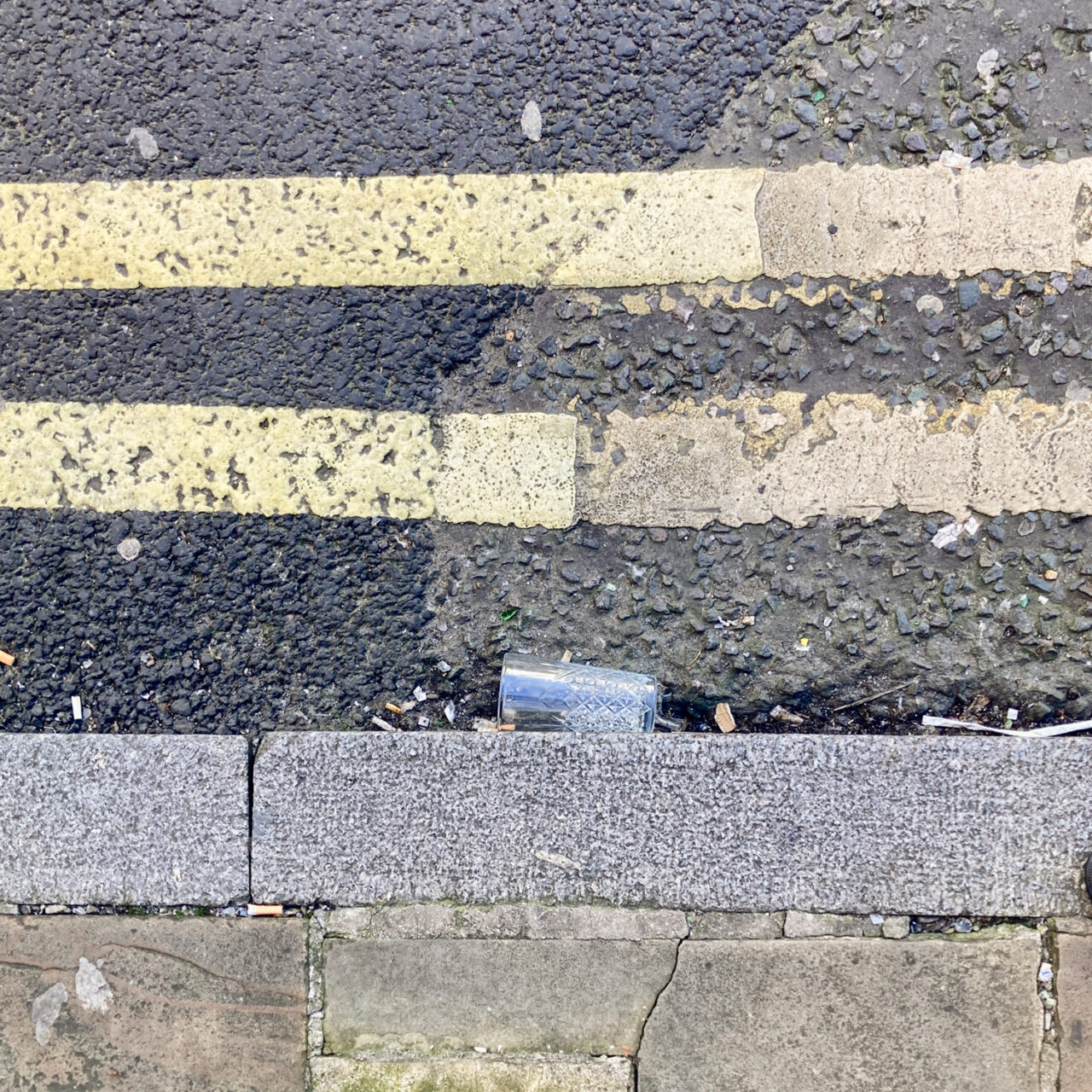 Photograph looking down at the gutter at the side of the road with the curb at the bottom of the frame, painted double yellow lines at the top of the frame and a cut-glass style highball glass on its side in the centre left of the frame.