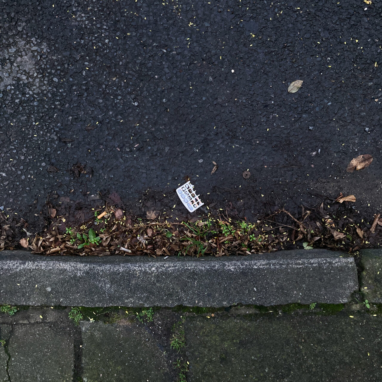 Photograph looking down at the curb at the side of a road, with pavement at the lower part of the frame, road above that and a piece of broken calculator in the gutter, road filling the top portion of the frame.