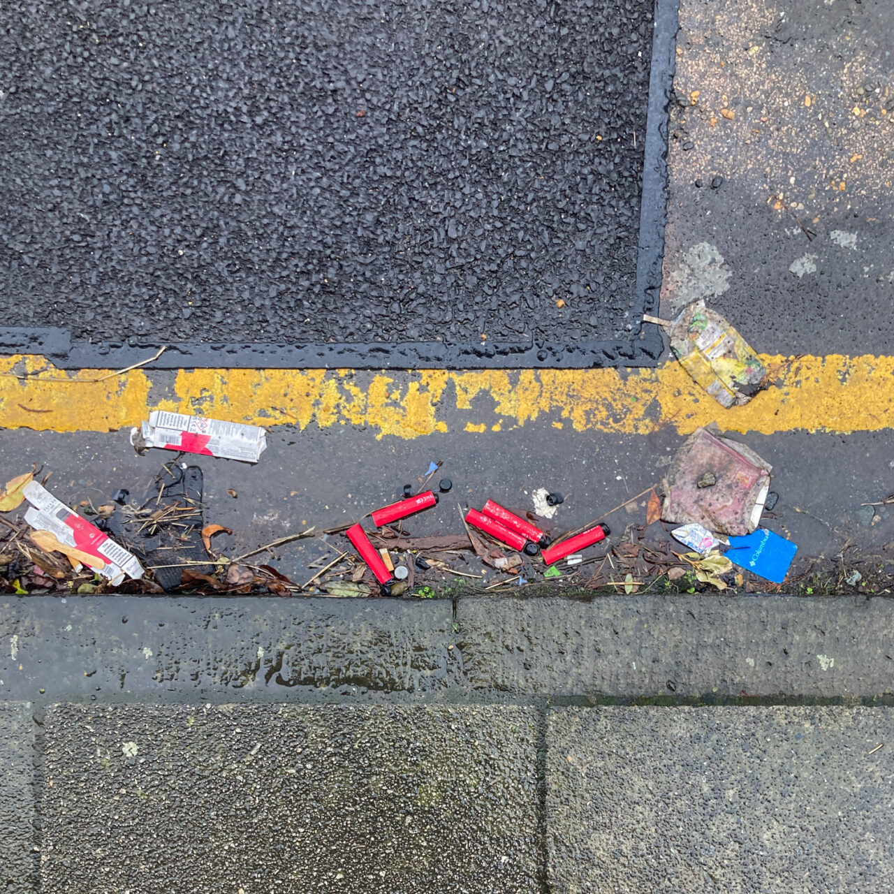 Photograph looking down at the curb at the side of a road, with pavement at the lower part of the frame, road above that and 5 red disposable vapes and packaging in the gutter, with a single yellow line above that and darker tarmac patching up the road filling the rest of the frame above.