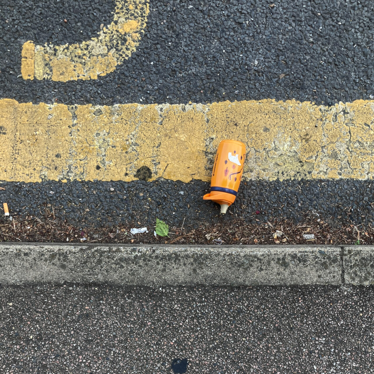 Photograph looking down at the curb at the side of a road, with pavement at the lower part of the frame, road above that and an orange plastic reusable drinks bottle in the gutter, with a thick single yellow line above that and a curved yellow painted line towards the top left.