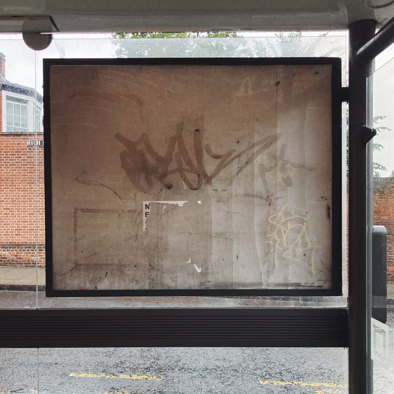 Photograph of an empty noticeboard in a bus shelter, with street behind and dirty marks and faded graffiti on it.