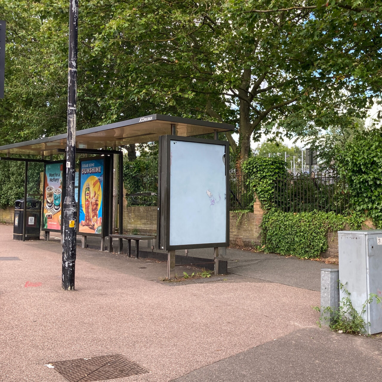 Photograph of an empty noticeboard on the side of a bus shelter, with street paving around it, trees and foliage behind, lampost, posters and a bin towards the left and a utility cover and grey metal utility boxes in the foreground.