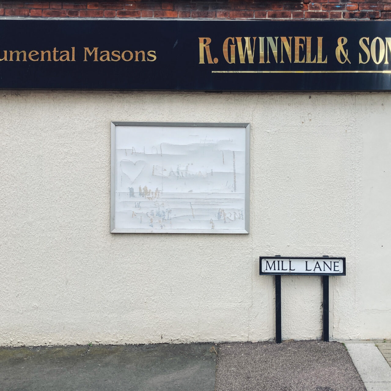 Walton-on-the-Naze, UK. Photograph of an empty noticeboard on a wall with a street name 'MILL LANE' to the right, '...umental Masons, R. GWINNELL & SONS' on a sign above and windows and brick wall above that, with pavement and double yellow lines at the bottom of the frame.