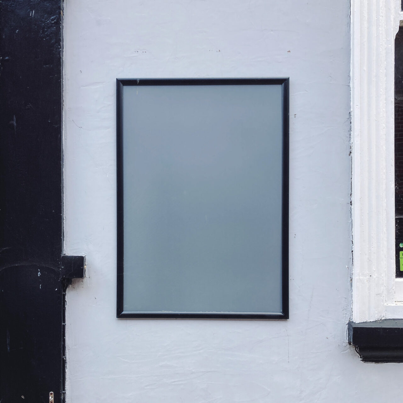 Photograph of an empty grey and black framed noticeboard on a light grey wall with black painted strip to the left and white painted windowframe just visible to the right.