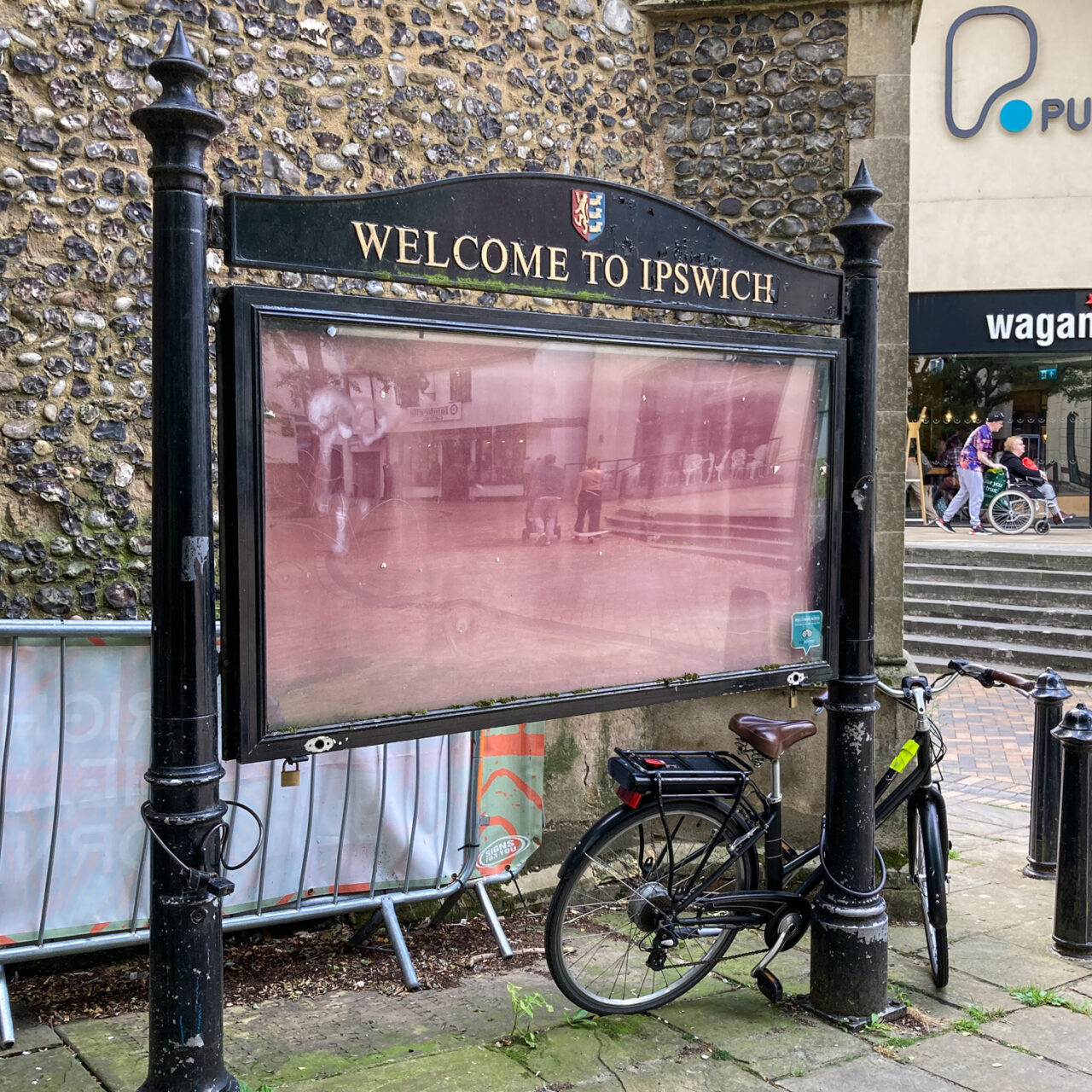 Photograph of a large metal noticeboard that says 'WELCOME TO IPSWICH' at the top with a coat of arms above and a faded red board under plastic below, which is reflecting bits of street scene in it. Locked to the right of the frame is a bike, there's a barrier and church wall behind,  and in the distance to the right are people passing a wagamama, which is up some stairs.