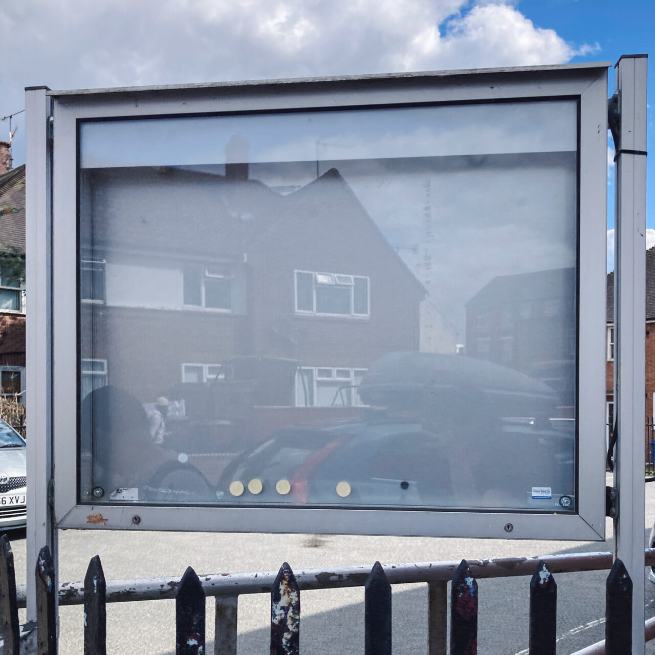 Photograph of an empty grey and metal framed noticeboard with a few circular magnets at the bottom, a reflection of houses and cars visible on the glass, sky with clouds visible behind above it and a metal fence below.