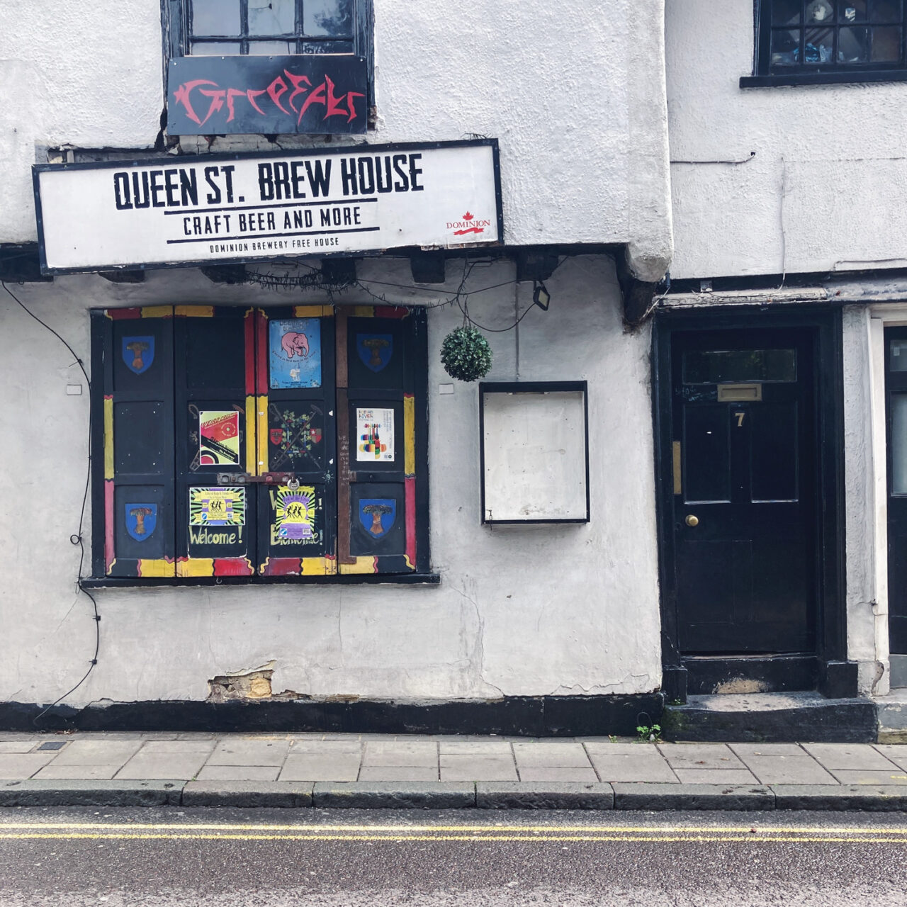 Photograph of a white painted old building with black door with '7' on it to the right, an empty black-framed noticeboard towards the centre, closed shutters with colourful paining and posters towards the left, a sign saying 'QUEEN ST. BREW HOUSE, CRAFT BEER AND MORE' above, pavement and road with double yellow lines in front at the bottom of the frame.