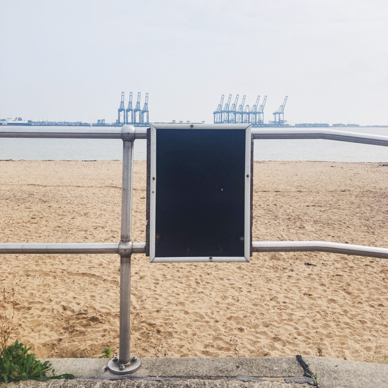 Photograph of an empty black noticboard with metal frame attached to a metal railing on a concrete floor, with sandy beach, sea and cranes at Felixstowe visible behind.