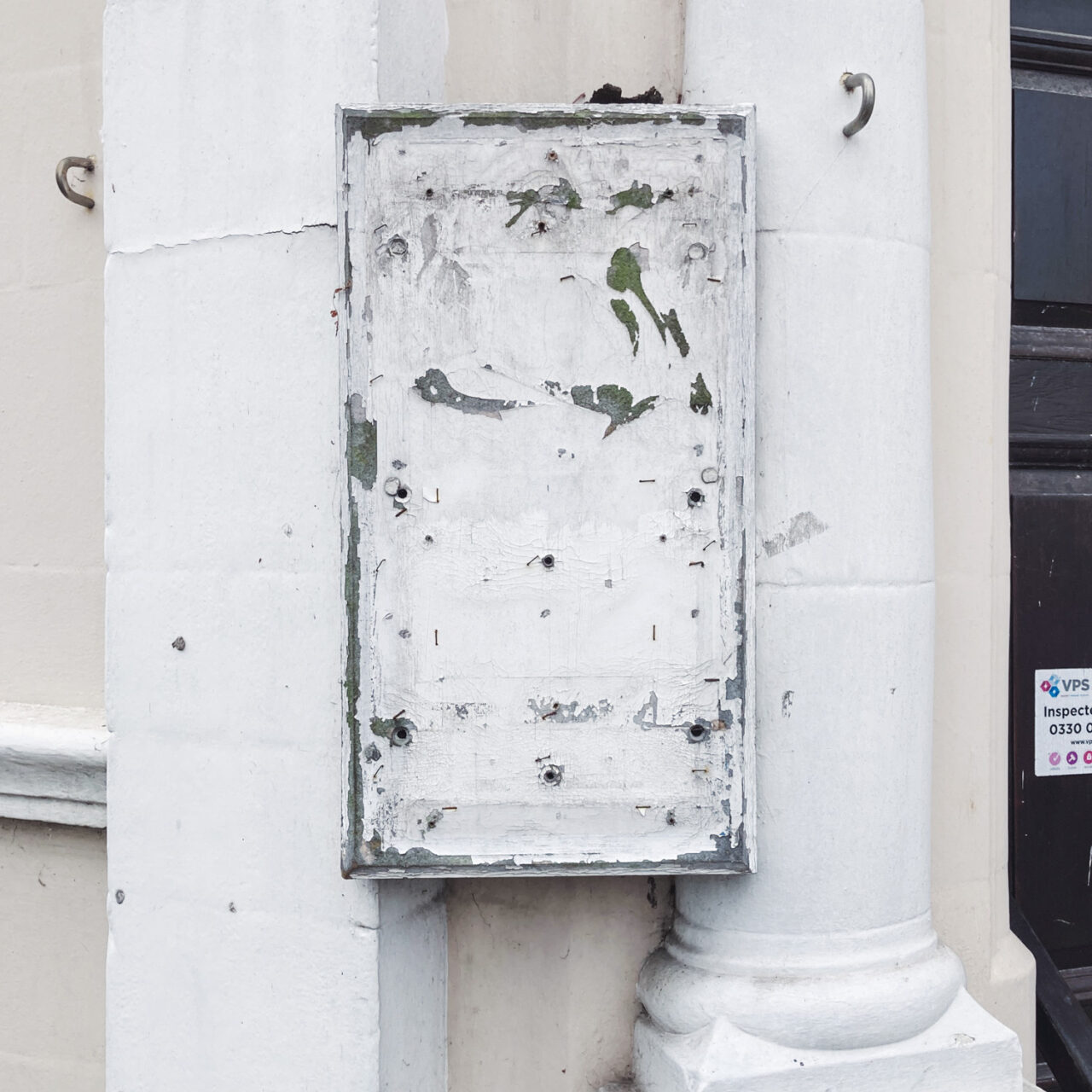 Photograph of a worn, white-painted noticeboard/sign with old staples on it, mounted on white columns against a cream building.