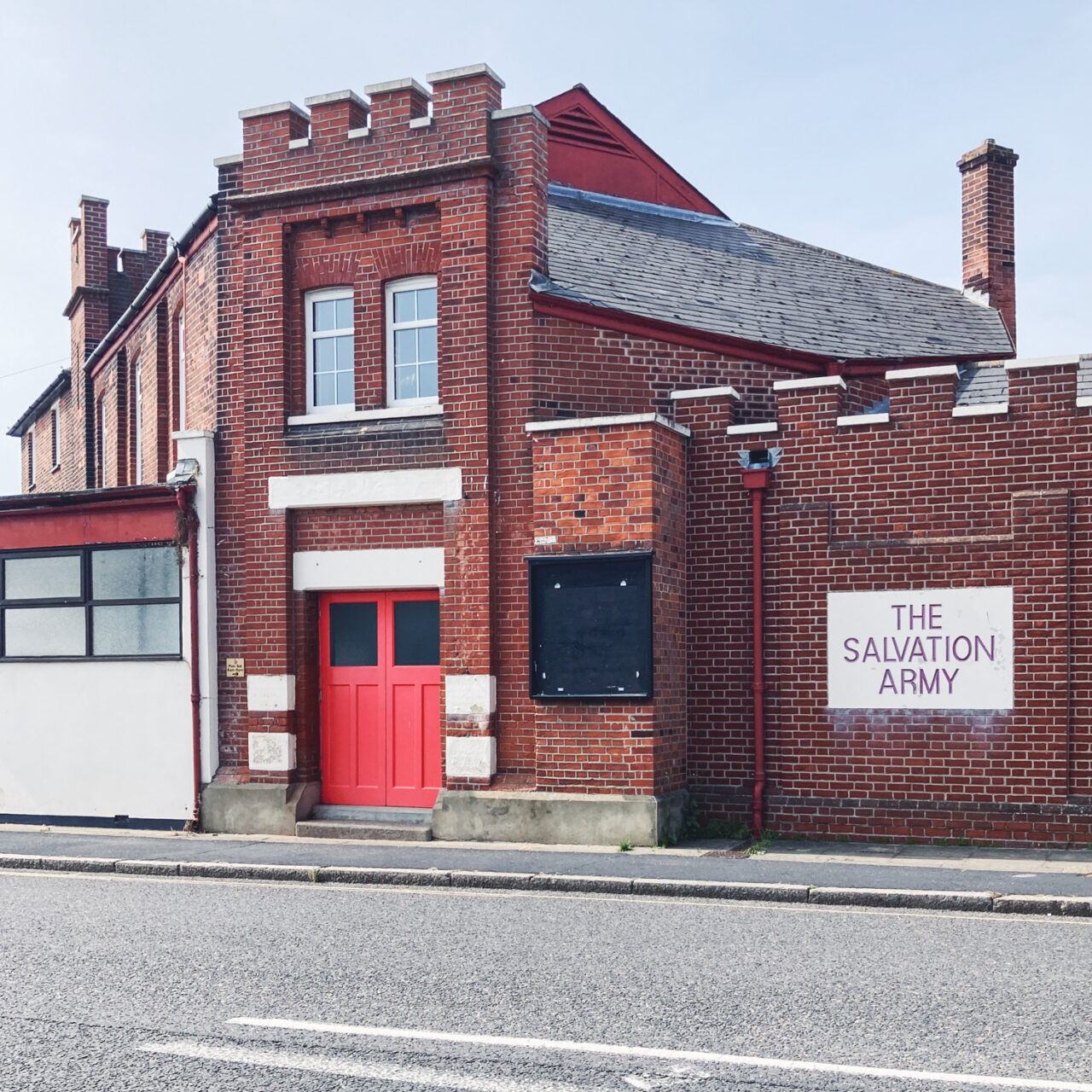 Photograph of a red brick building with turret style walls, a painted sign saying 'THE SALVATION ARMY' to the right, a large, black-painted empty noticeboard towards the centre, a red double door towards the left and windows above and to the left of frame. Pavements and road are visible in the foreground.