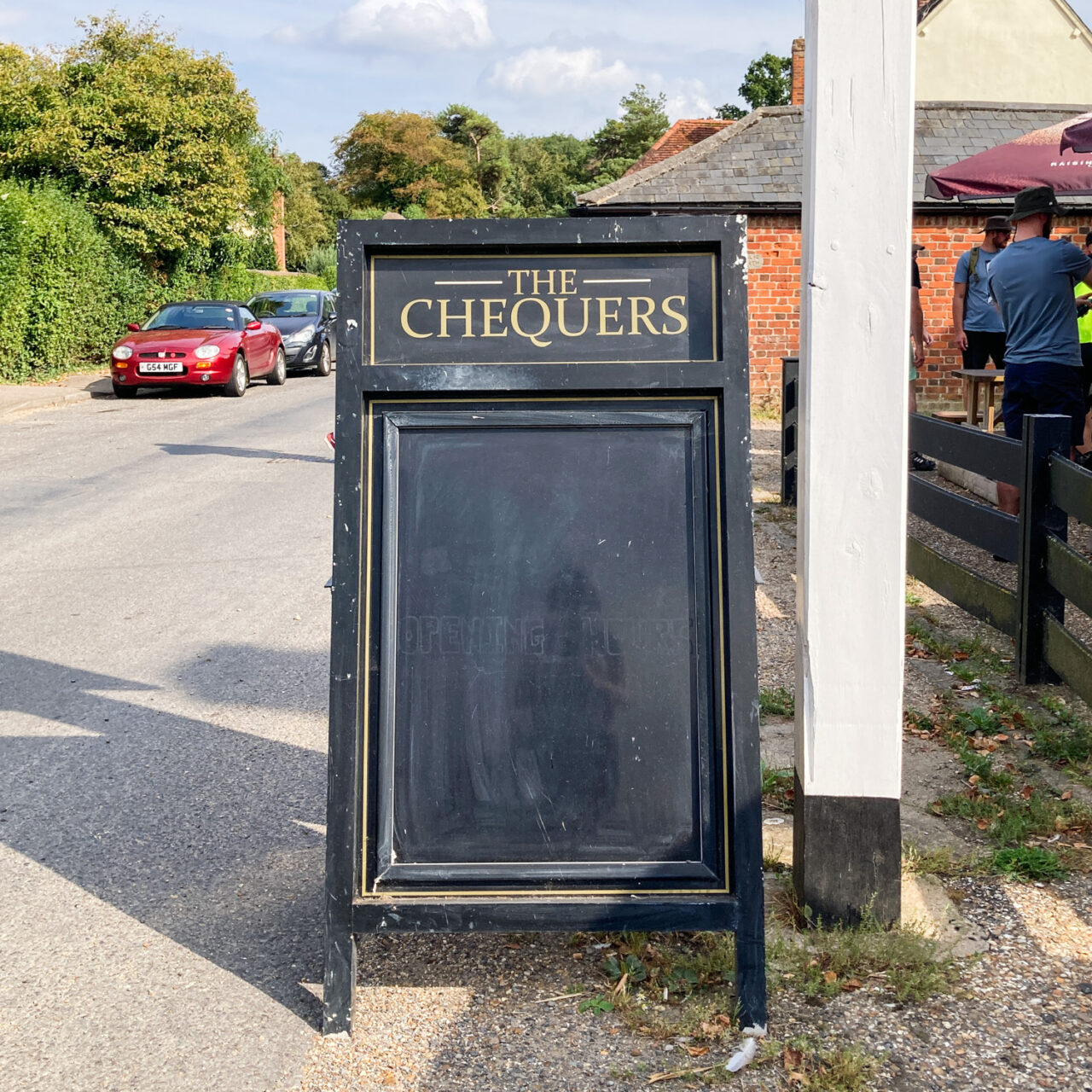 Photograph of a wooden A-frame sign that has 'THE CHEQUERS' writeen at the top and black chalkboard below. It has a white wooden pole to the right of the frame and people behind a fence further to the right, a road with cars parked along the edge and trees to the left.