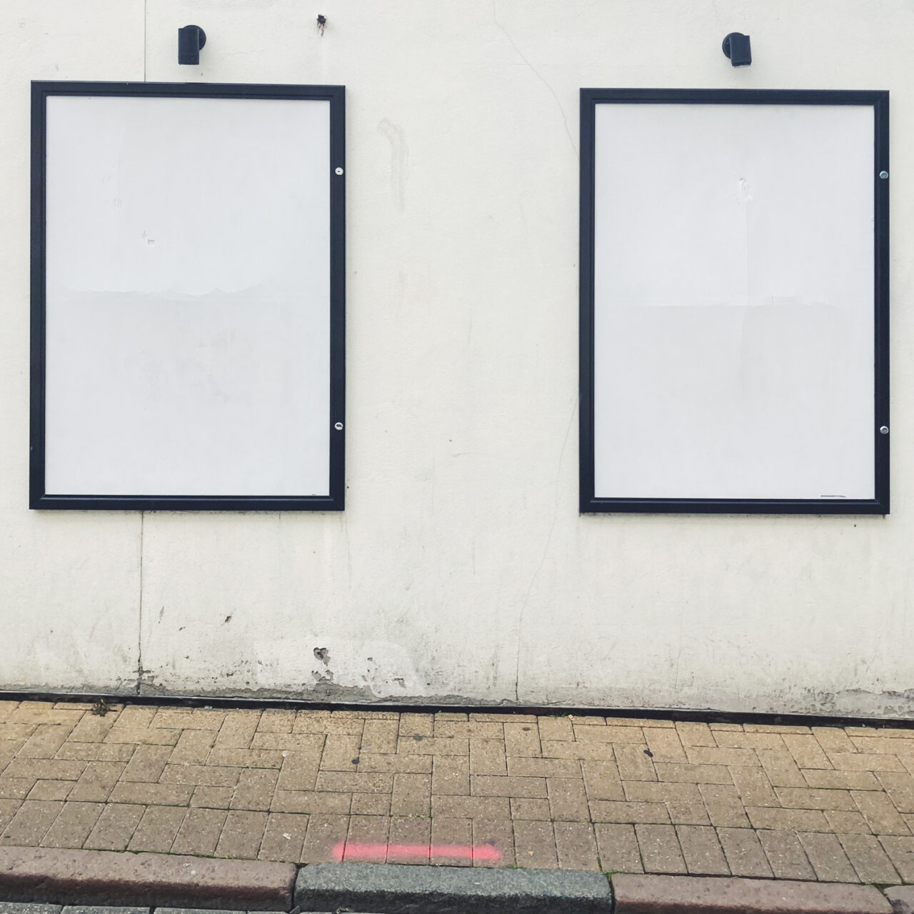 Photograph of two empty white noticeboards with black frames on a white wall, with small black lights above them, with brick pavement and curb below and towards the bottom of the frame.