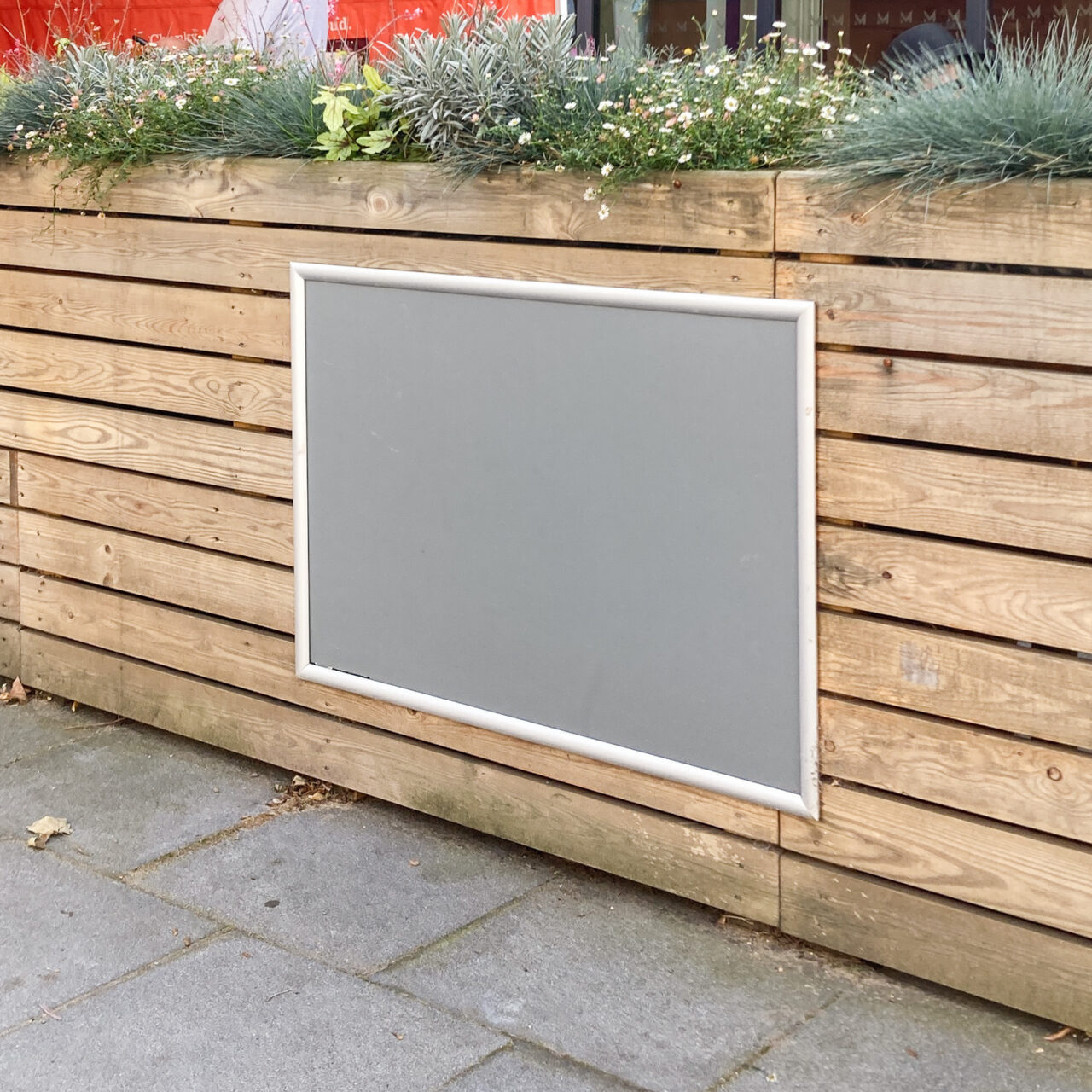 Photograph of an empty grey and metal framed noticeboard on a wooden planter with plants visible at the top of the fram and pavement visible in the foreground.