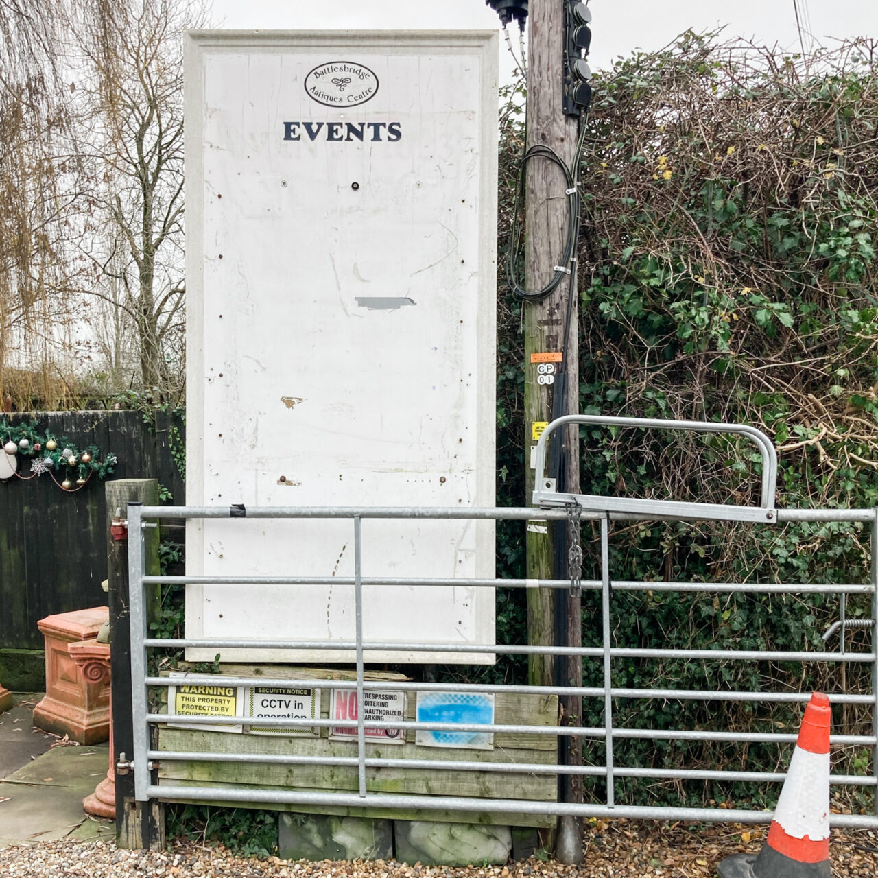 Photograph of a large empty white board with the Battlesbridge Antiques Centre logo at the top and 'EVENTS' below, with only drawing pins and scratches below. The board is behind a metal gate, there are CCTV warning notices below, terracotta pots to the left, trees and plants behind and a traffic cone to the bottom right.