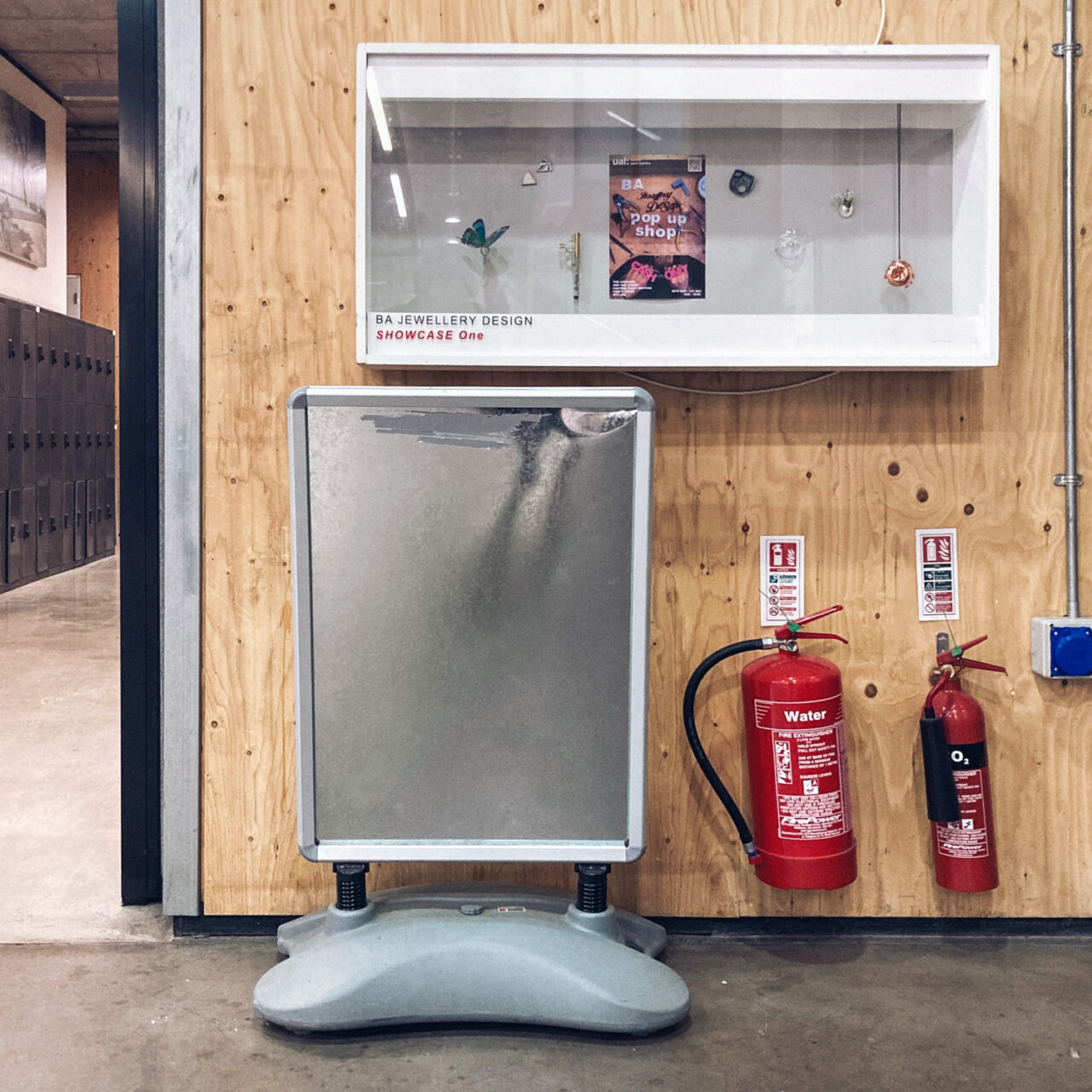 Photograph of a free-standing metal noticeboard with a grey plastic base and nothing displayed on it. Above it, mounted on a bare wooden wall is a display case with objects on display and 'BA JEWELLERY DESIGN, SHOWCASE One' written at the bottom. Below that are two fire extinguishers, there's a concrete floor below and at the left of the frame, in the distance, are grey metal lockers.