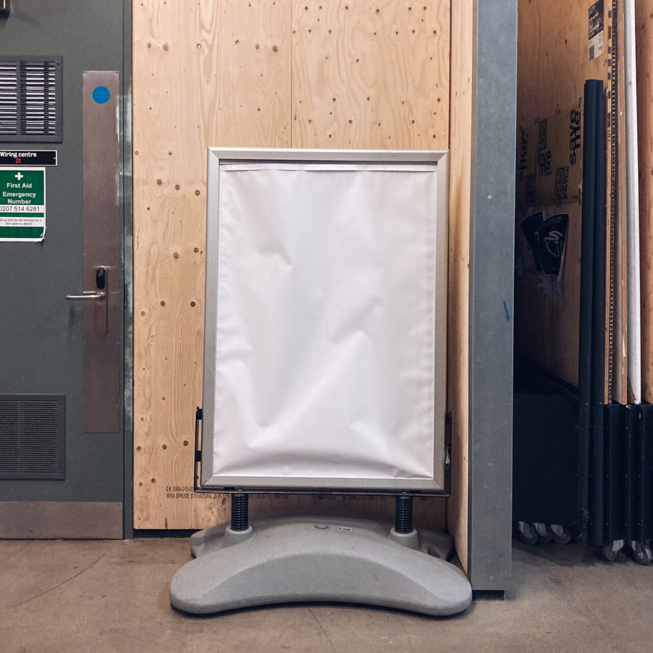 Photograph of a free-standing  noticeboard with a grey plastic base and a blank white sheet of paper displayed. Behind is a bare wooden wall, to the left is part of a door, to the right are boards stacked for storage and there's a concrete floor below.