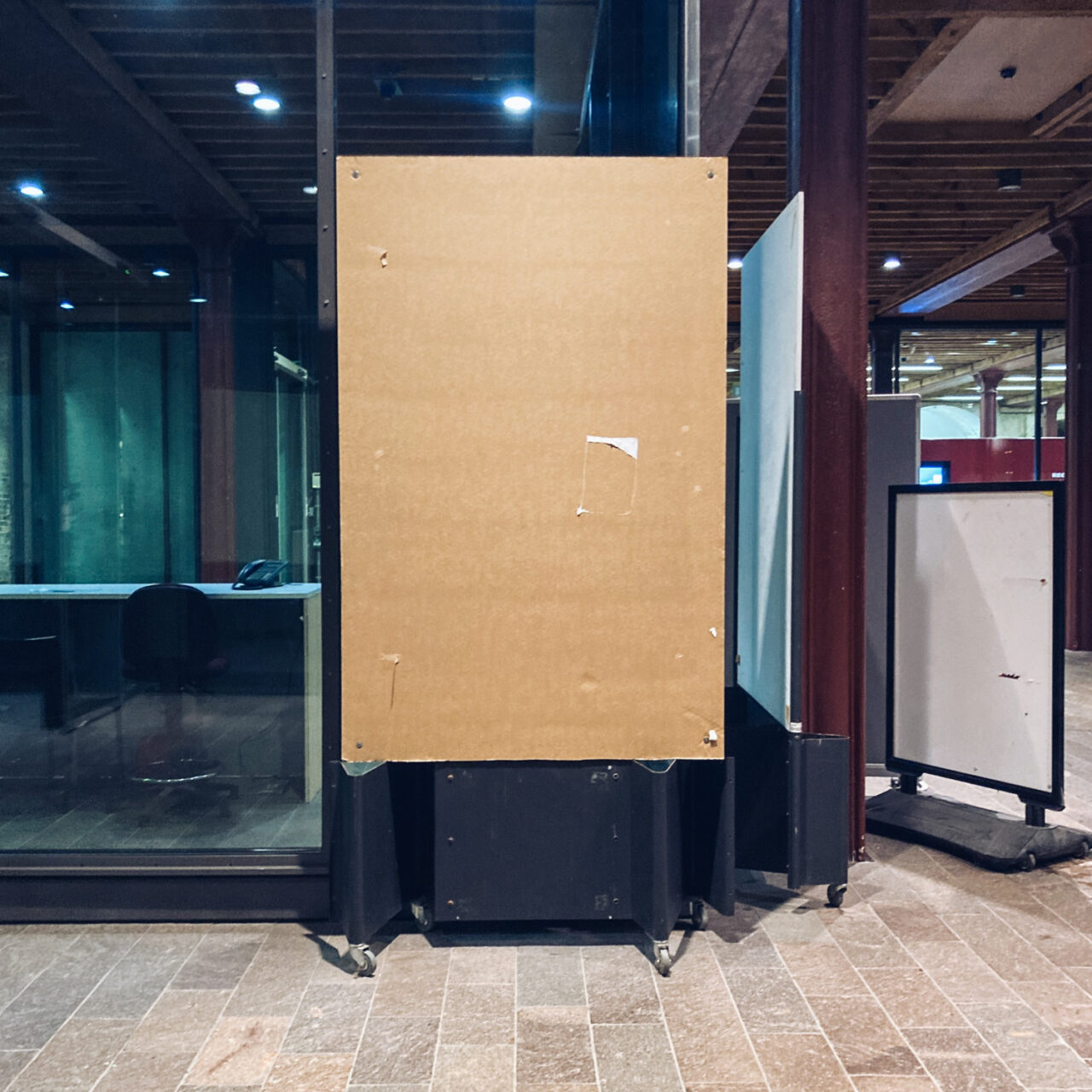 Photograph of a free-standing wooden noticeboard with nothing on it. To the right of the frame is another freestanding board with a blank white sheet on it, behind is a glass window into an office and at the bottom of the frame is a paved floor.