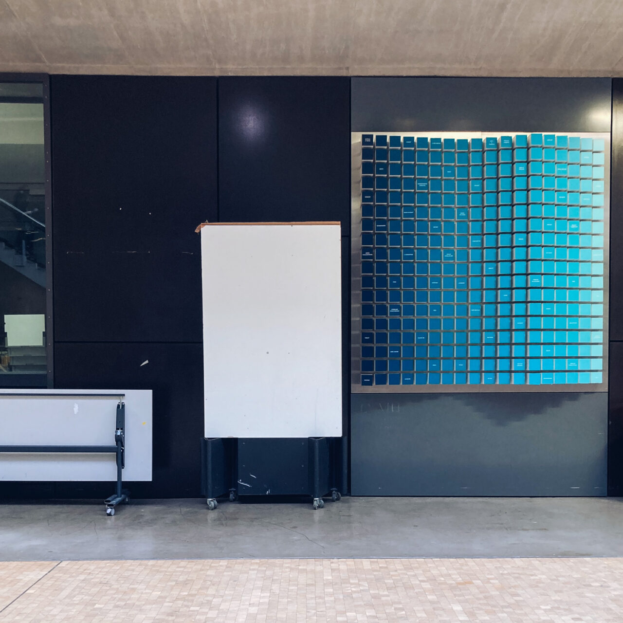 Photograph of a free-standing noticeboard with just a blank white board. behind is a black wall, to the right is a grey wall with a display of different blue toned squares, to the left is a folded up white table and in the foreground is a concrete floor and tiled floor closer to the foreground.