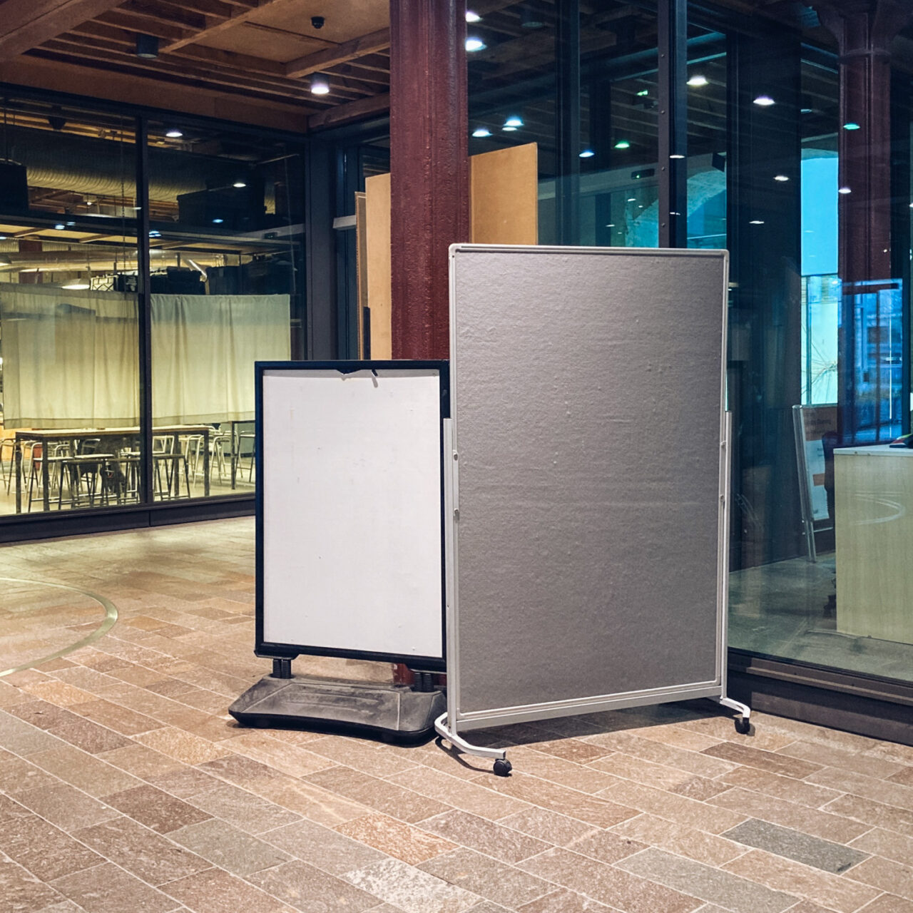 Photograph of two empty free-standing noticeboards in the entrance of a building, a white one to the left and grey to the right.