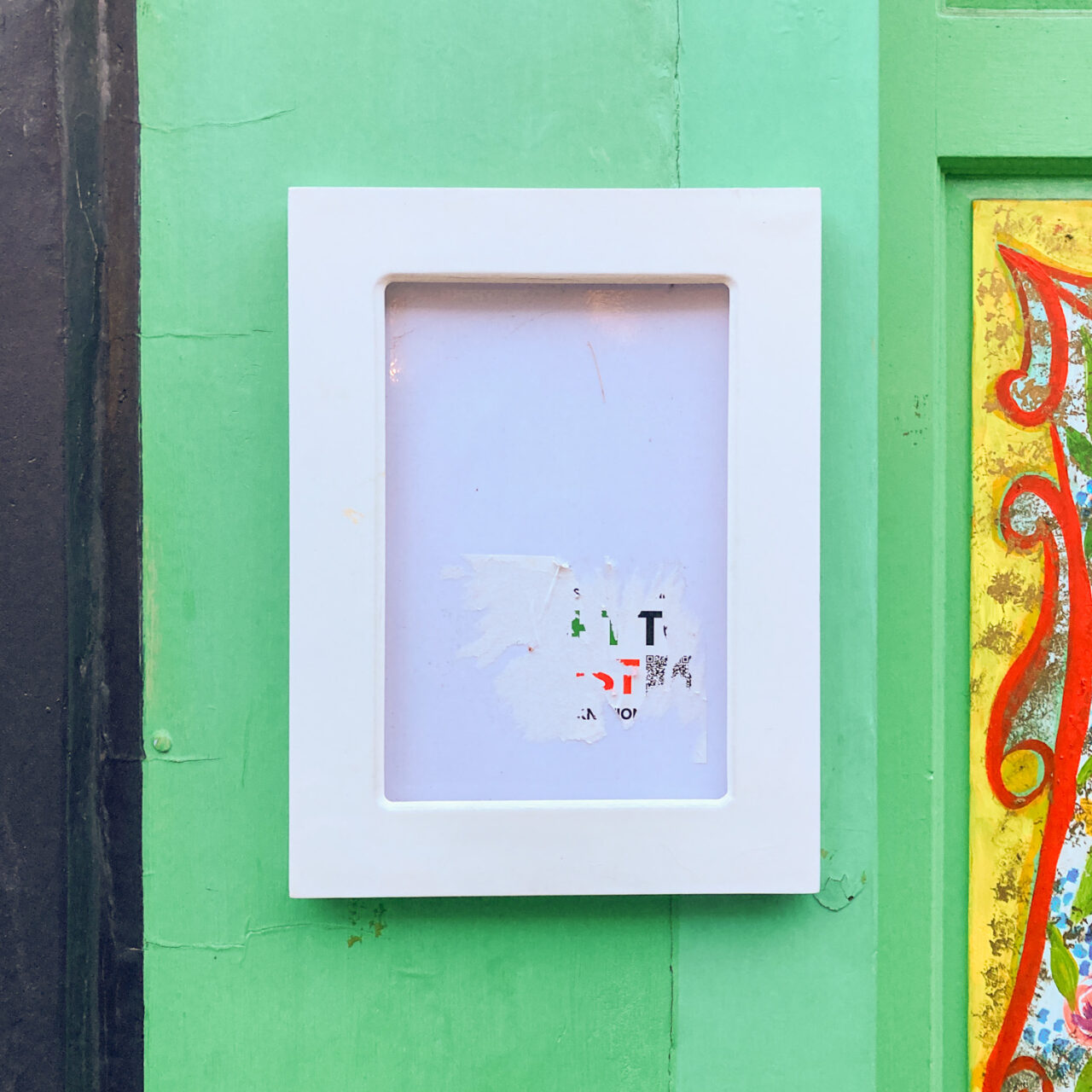 Photograph of a wide-edged white noticeboard with remnants of a sticker on the glass but empty inside. Behind is a bright green wall with a black strip to the left and a yellow and red pattern painted area to the right.