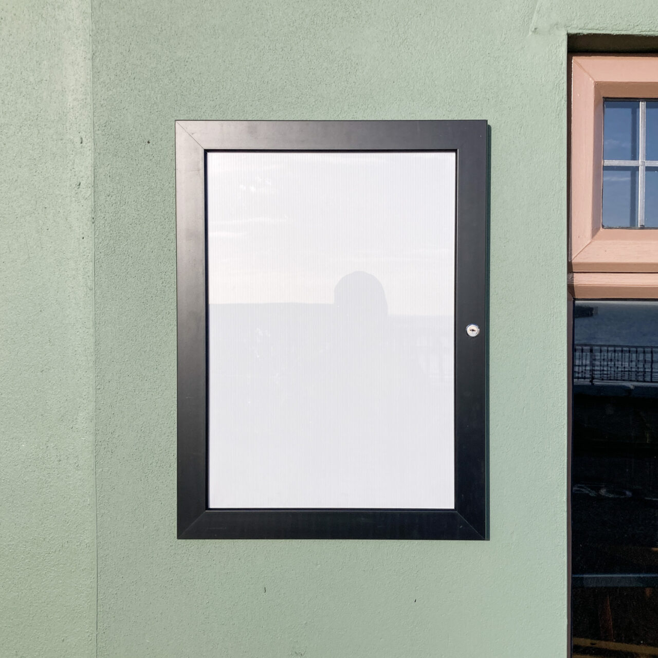 Photograph of a wide-edged black frame around an empty white noticeboard. Behind is a light sage-green wall, with part of a window visible to the right of the frame.