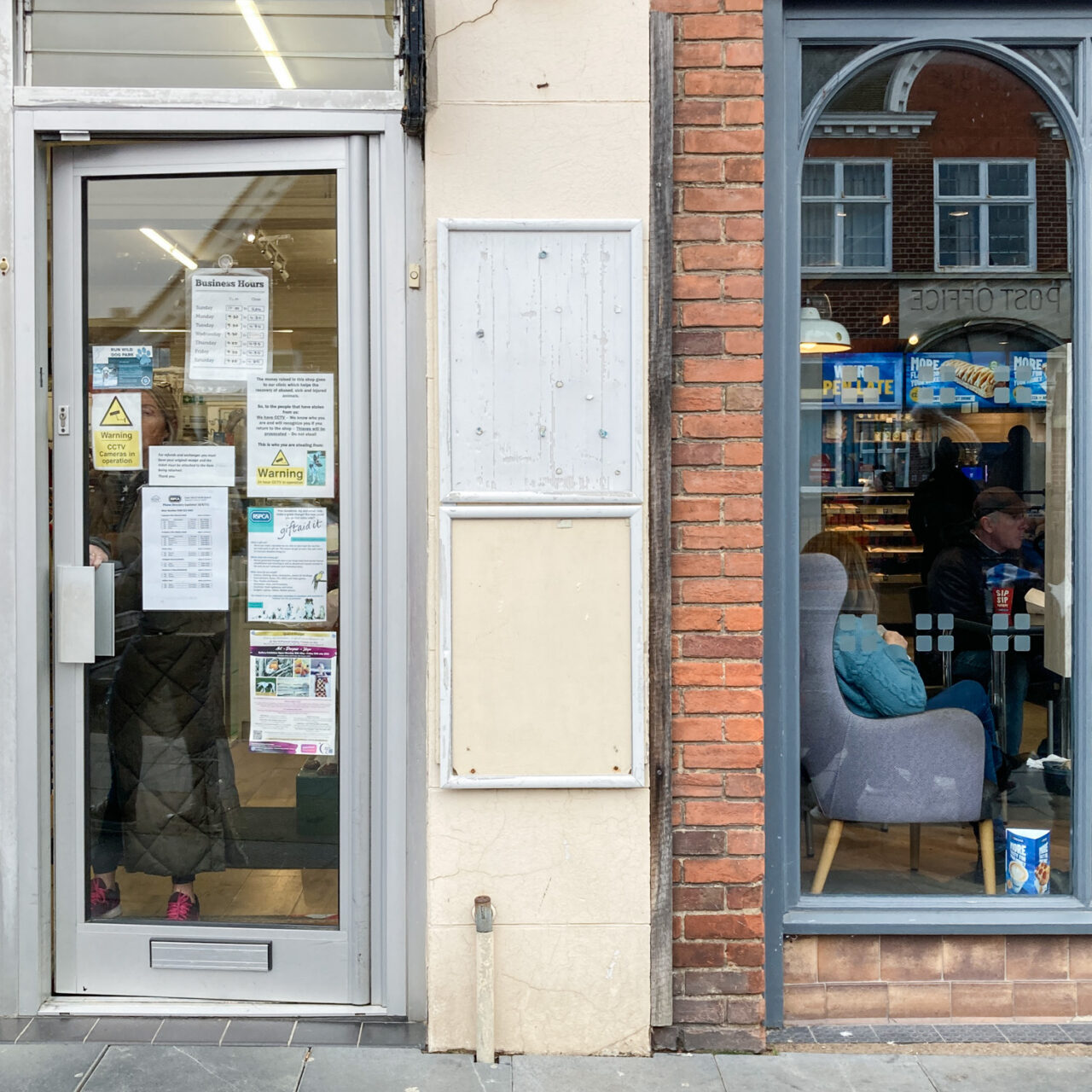 Photograph of two empty noticeboards mounted on a strip of cream wall, the top one with a worn white background and the lower just showing the wall behind. To the left is a metal and glass shop door with various signs showing Business Hours and Warning notices and to the right is a more ornate window into a cafe, with an armchair by it.