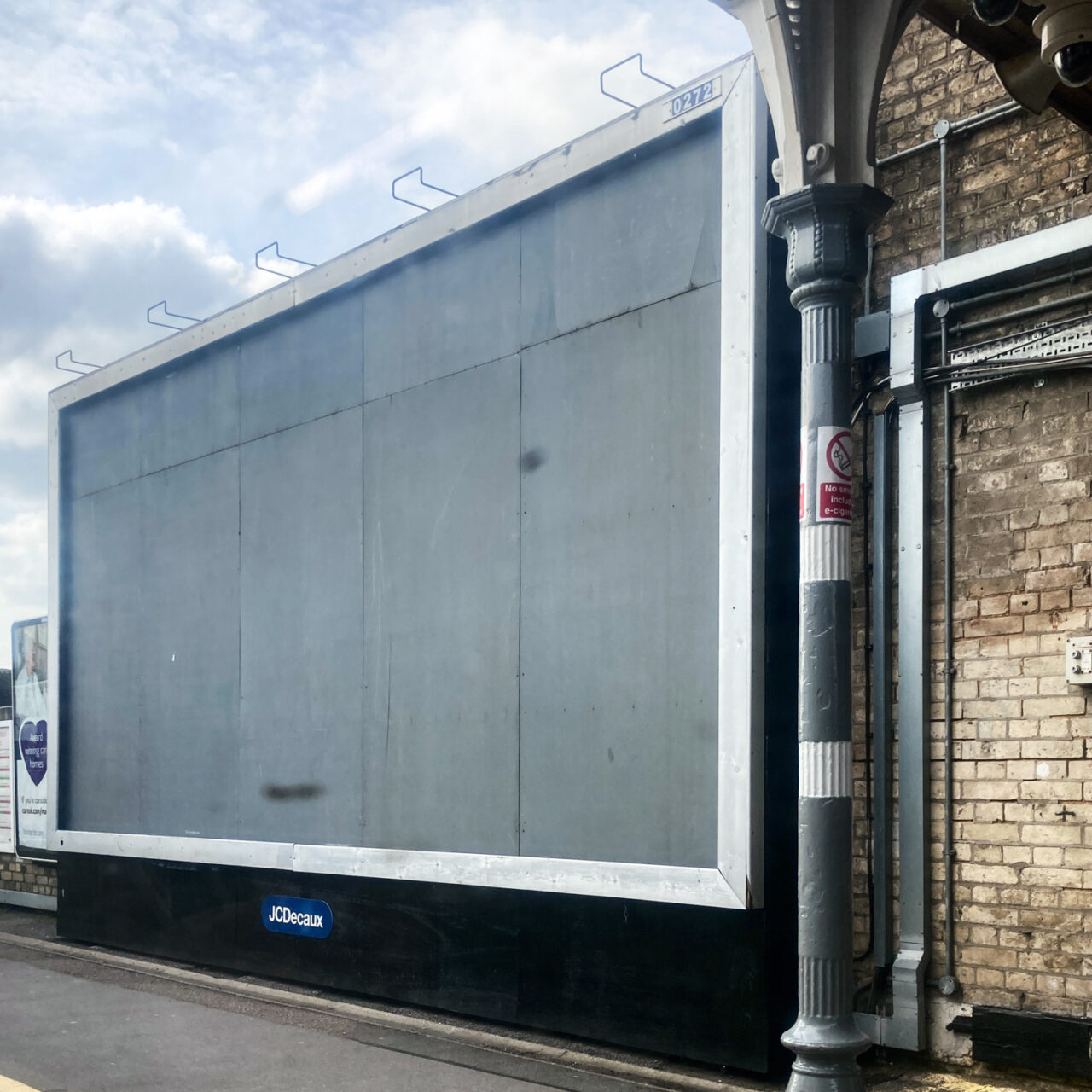 Photograph of a large advertising board on a train platform, viewed at angle. It's empty, with grey in it and a metal frame. Above and to the left is a cloudy sky and to the right is a pole, pipes and brick wall.