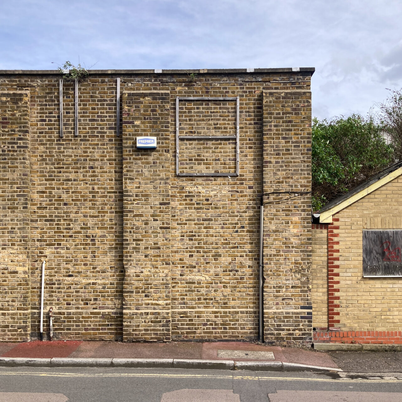 Photograph of a brick wall with an empty wooden frame towards the top. To the left of it is an alarm casing, with wooden batons to the left of that. To the right is a house-shaped lower brick wall, with a boarded up window. Above is a blue and cloudy sky and below is a pink pavement with double yellow lines on the road.