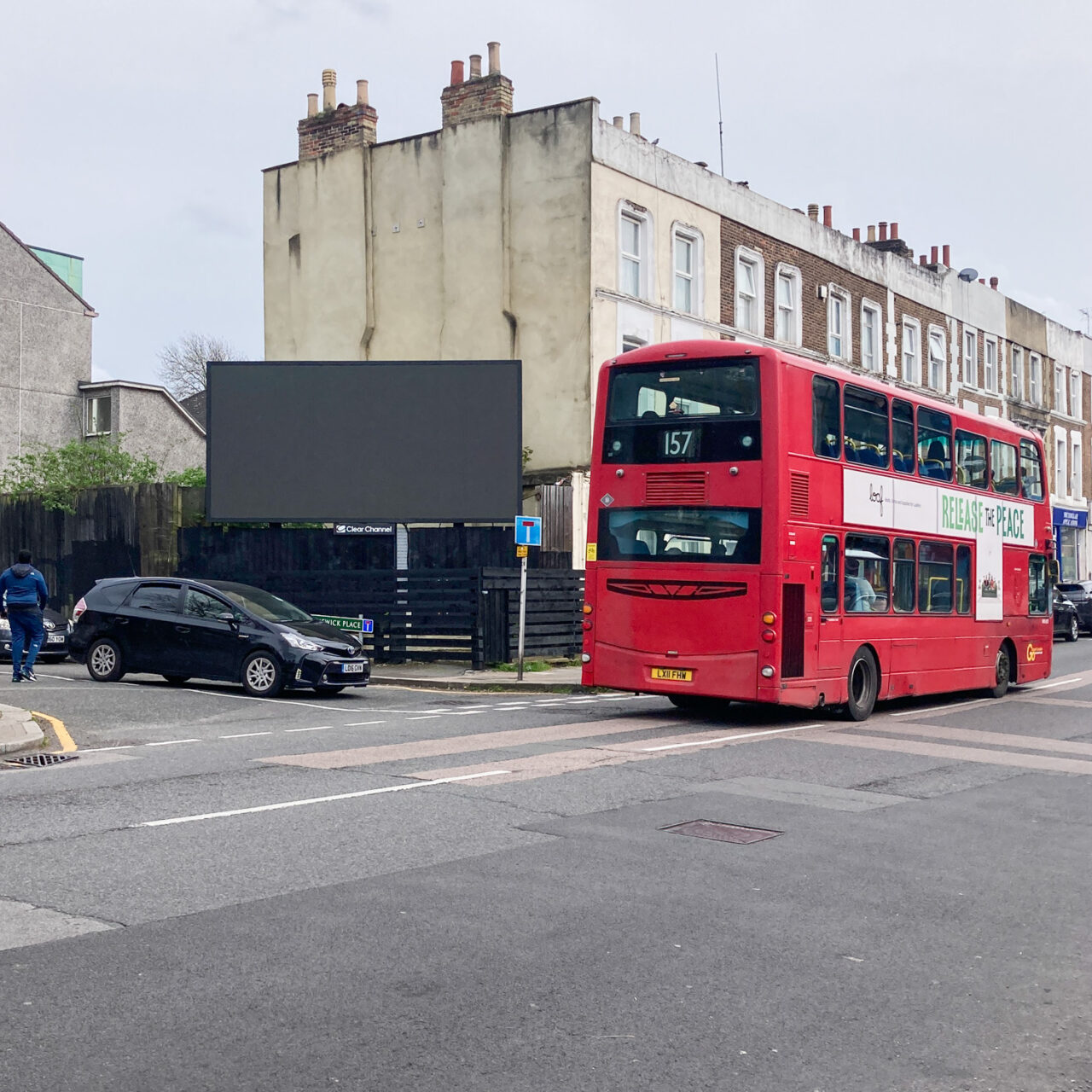 Photograph of a large advertising board on the corner of a road - it is empty and black. In front of it a black car is turning onto the main road, on which there is a red London bus. There is a row of houses behind and the foreground is filled with road.